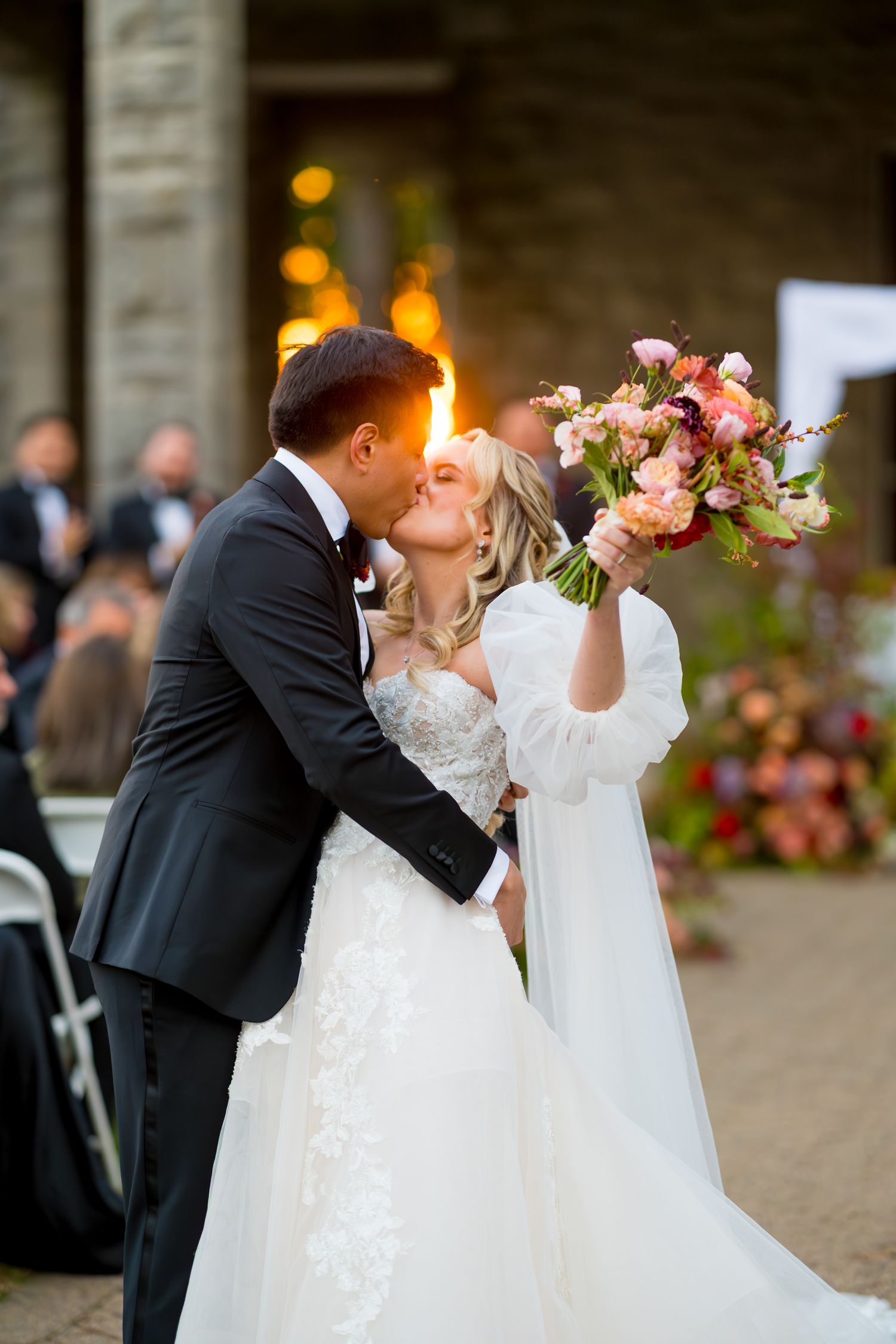 A romantic wedding couple shares a kiss outdoors with floral bouquets and guests in the background.
