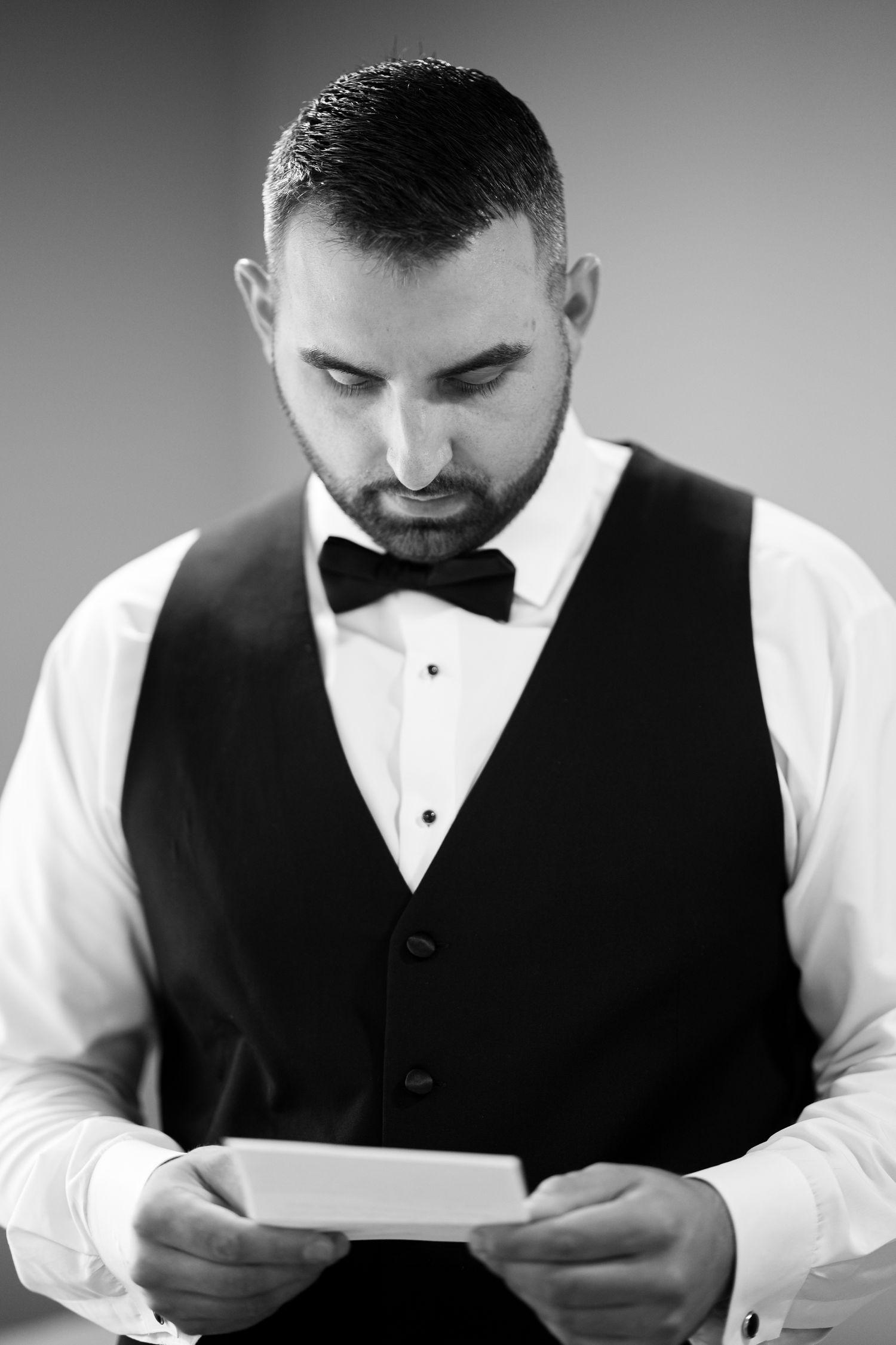 Man in formal vest and bow tie reading a document in black and white.