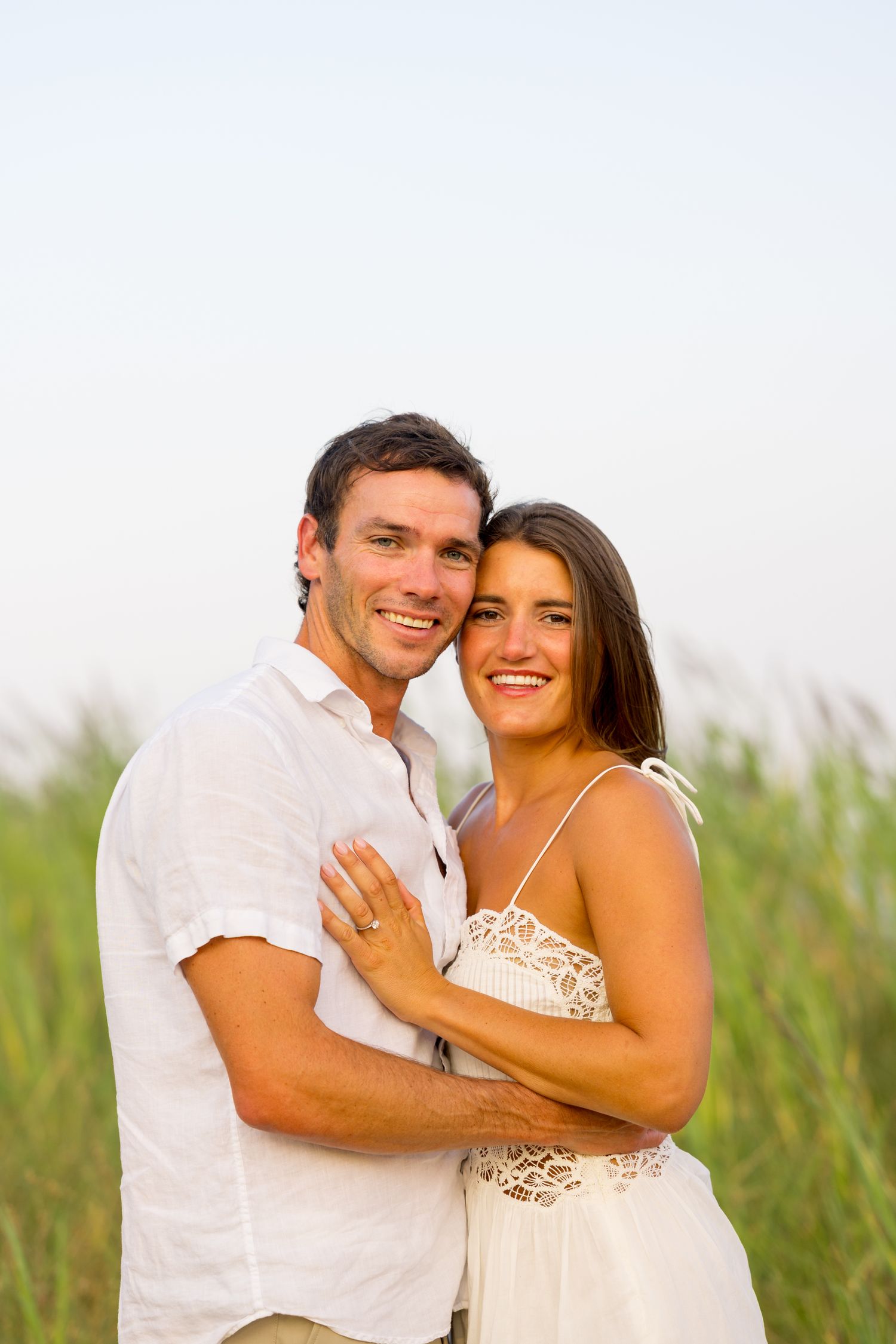 A couple in white outfits embrace and laugh together in a field of tall grass during a sunny outdoor photo session.