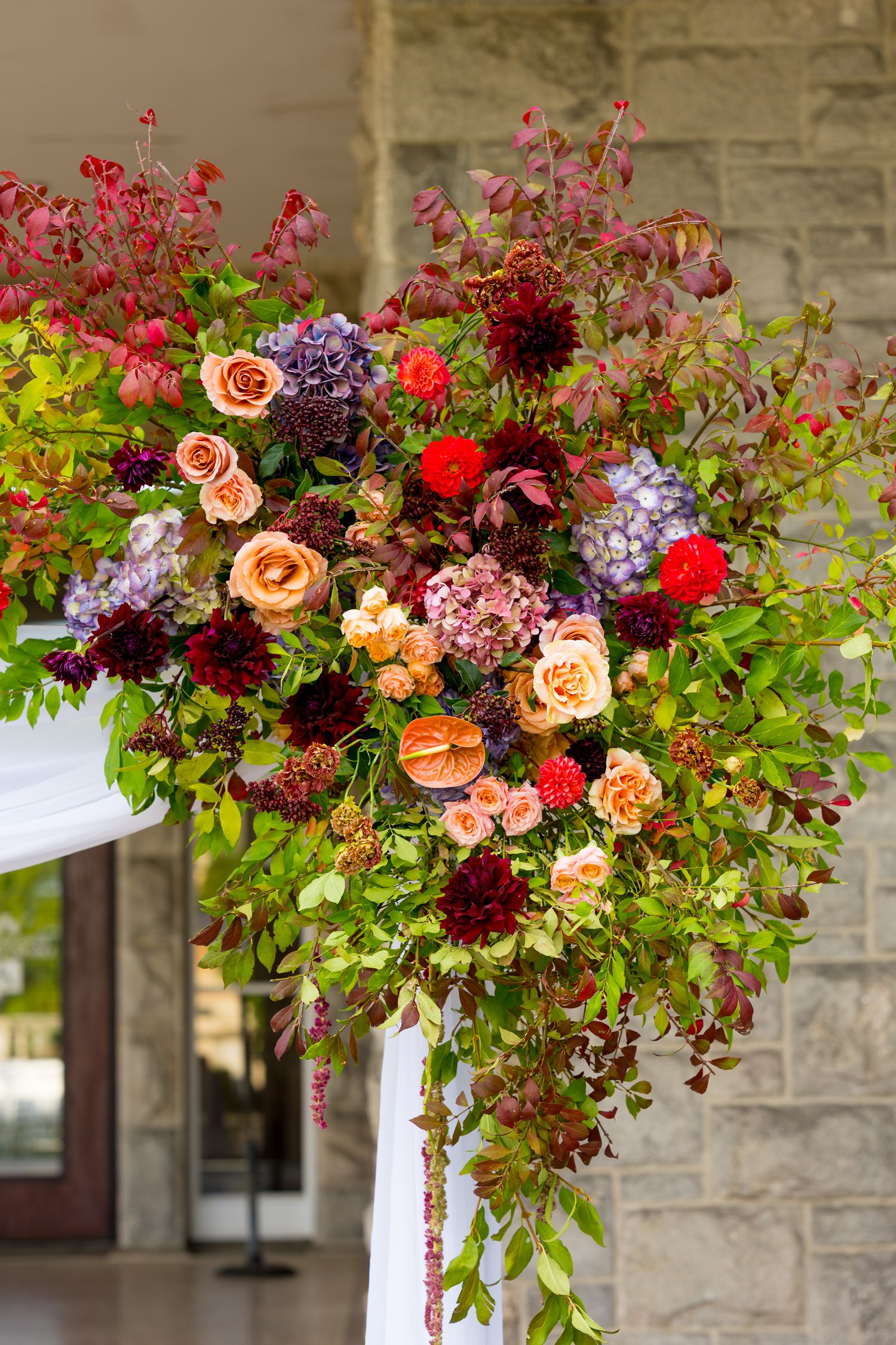 Lush floral arch installation featuring peach roses, red blooms, and purple hydrangeas against a stone wall backdrop.