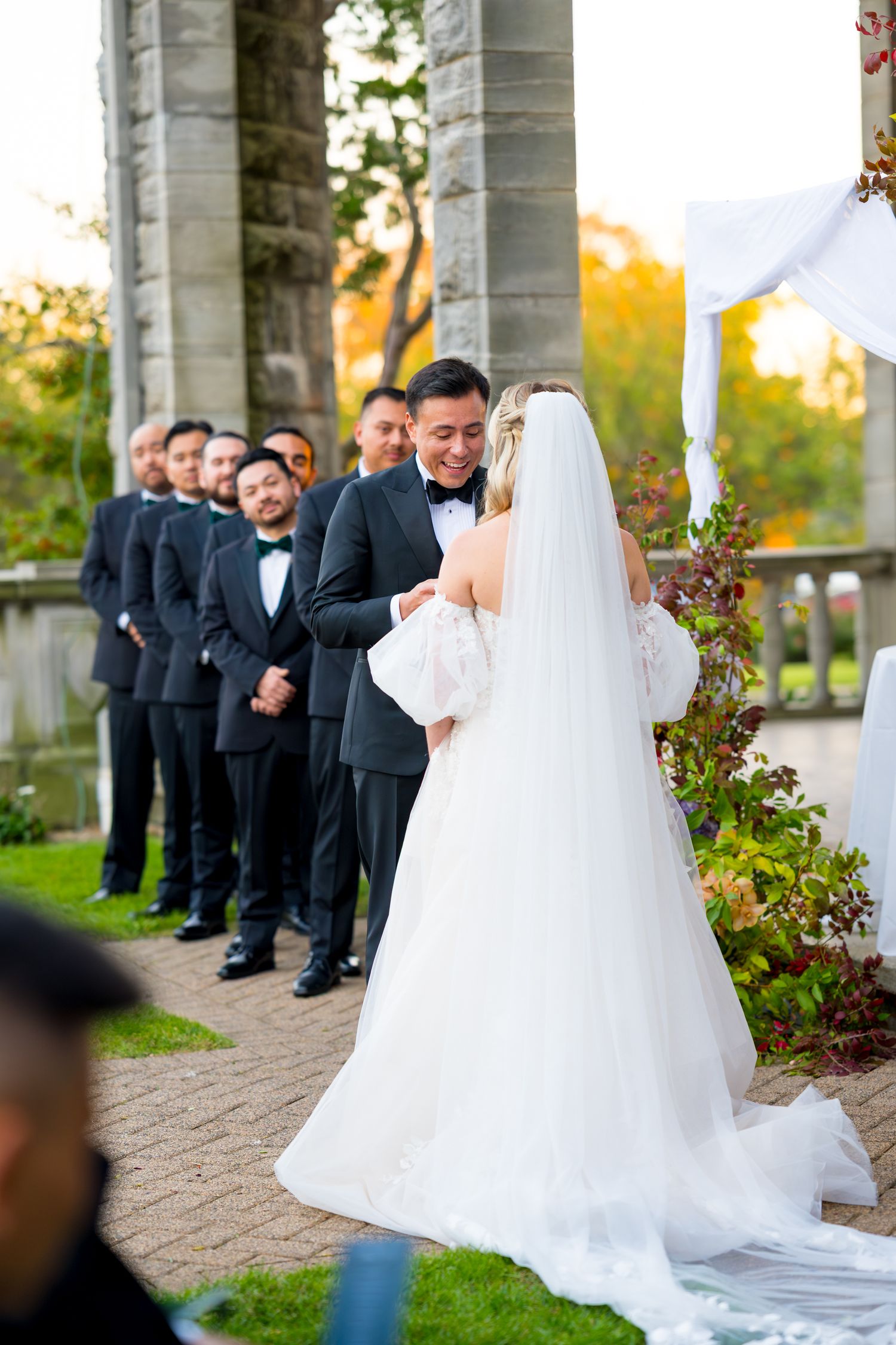 Wedding party stands in line during outdoor ceremony as bride in flowing white gown and veil faces groomsmen.