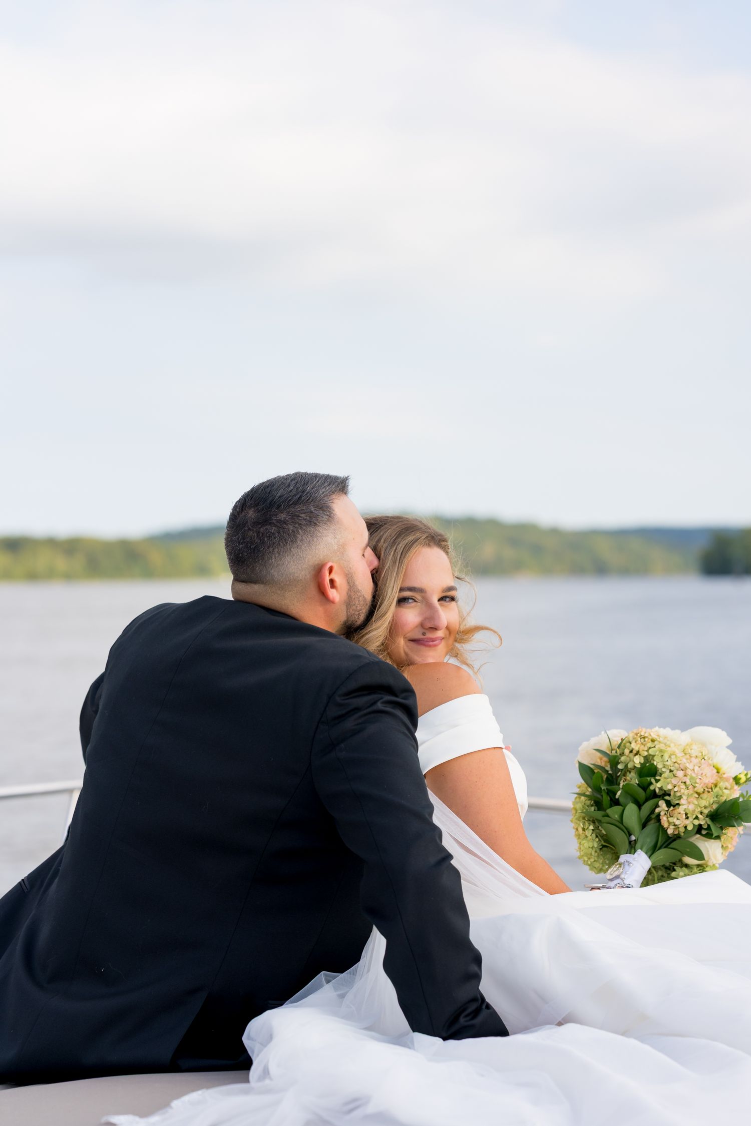 Romantic couple in wedding attire sitting together by a serene waterfront.