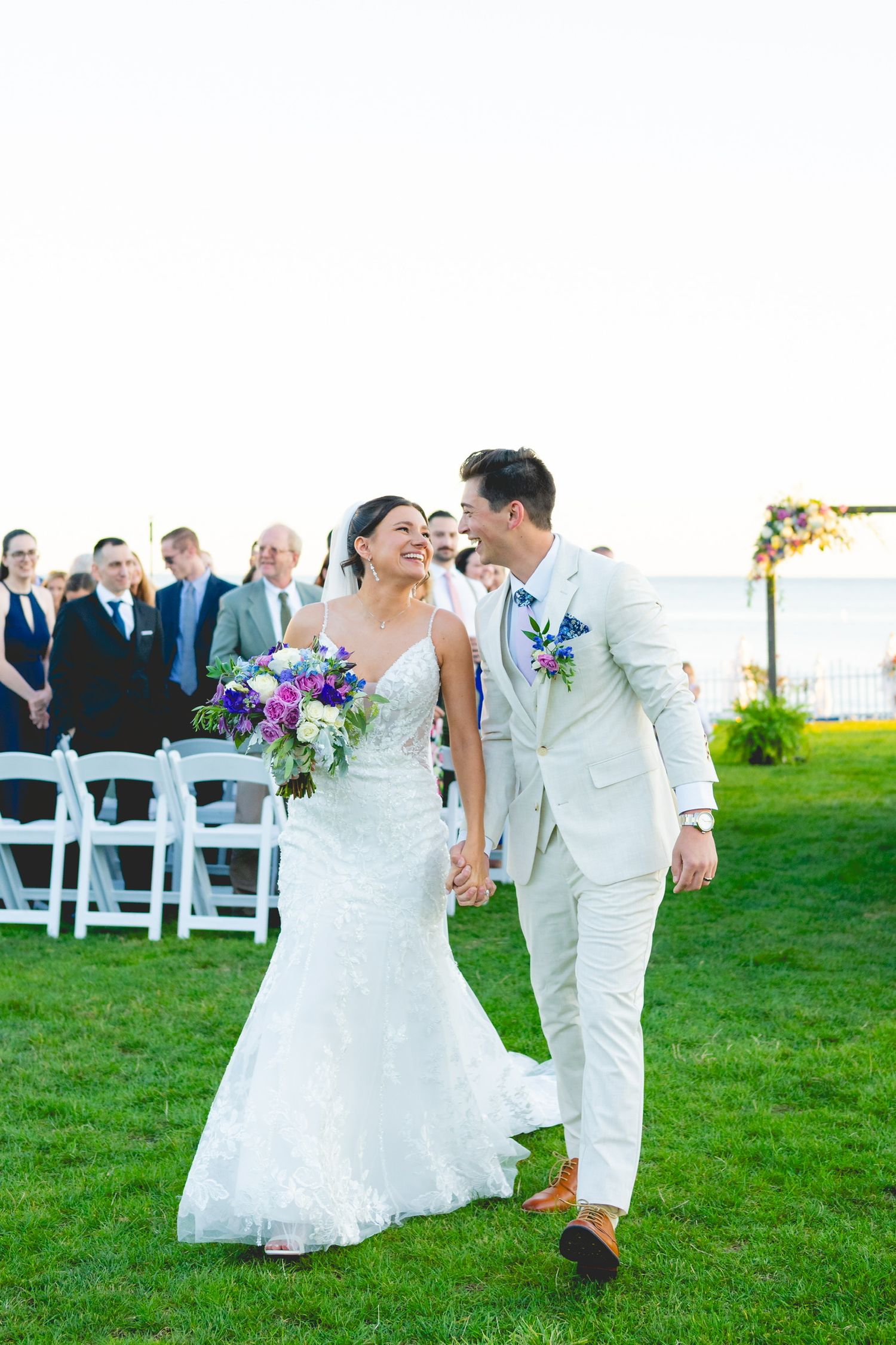 Newlyweds share a joyful moment walking on grass at their outdoor wedding ceremony with guests in background.