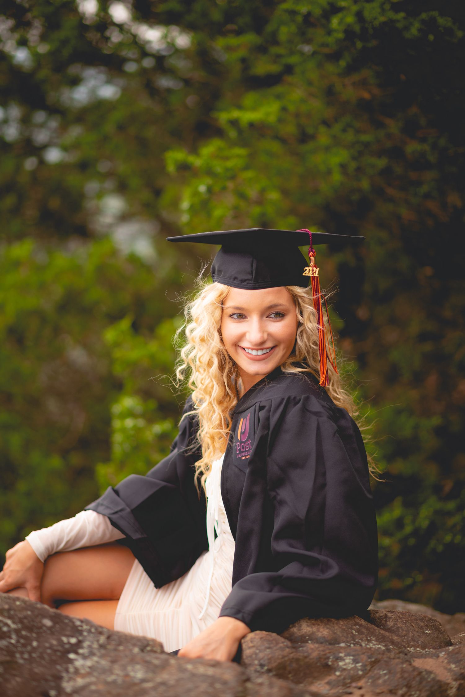 Graduate in black cap and gown poses outdoors for professional portraits in natural setting.