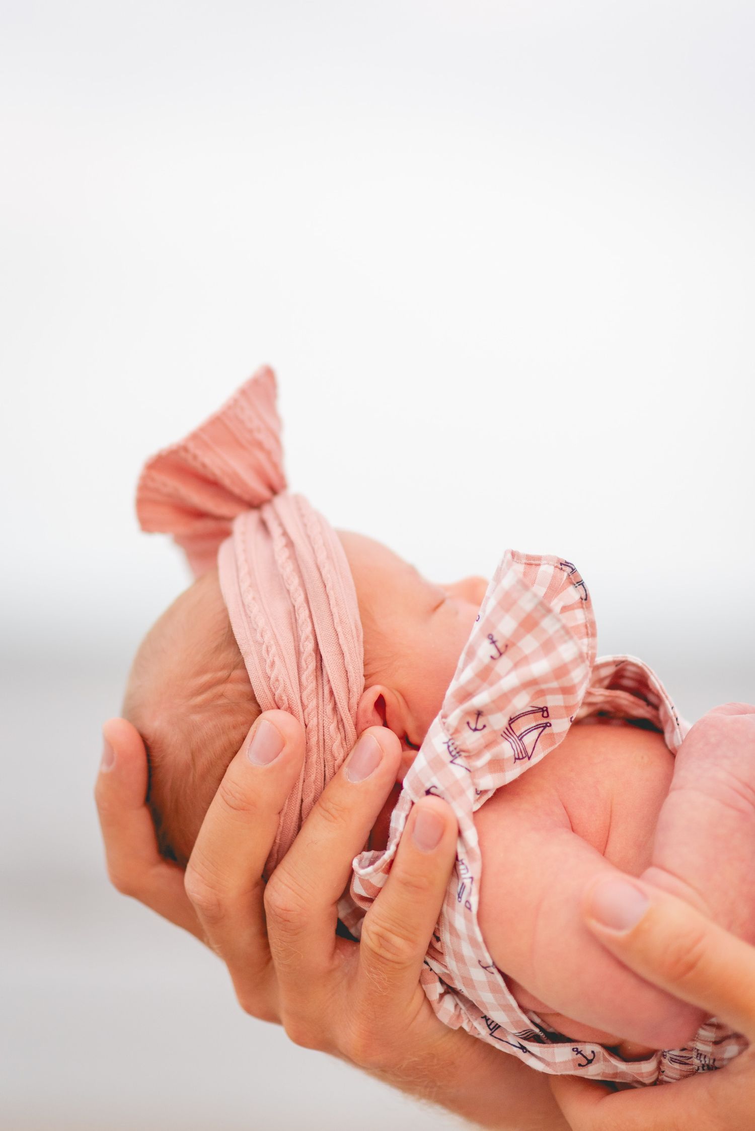 Close-up of hands gently holding a newborn baby wrapped in pink checkered fabric.