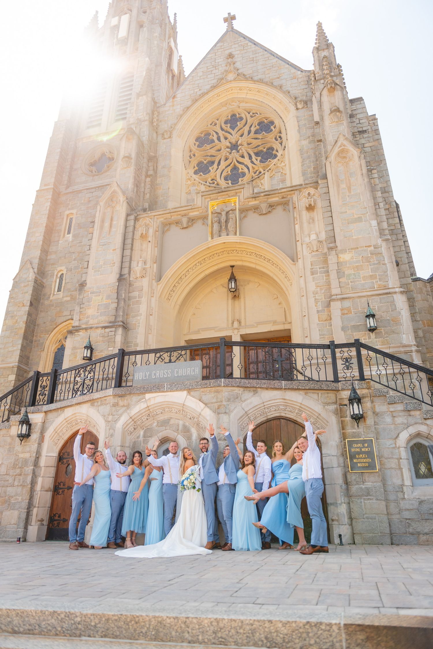 Wedding party poses on church steps with bridesmaids in light blue dresses against gothic architecture.