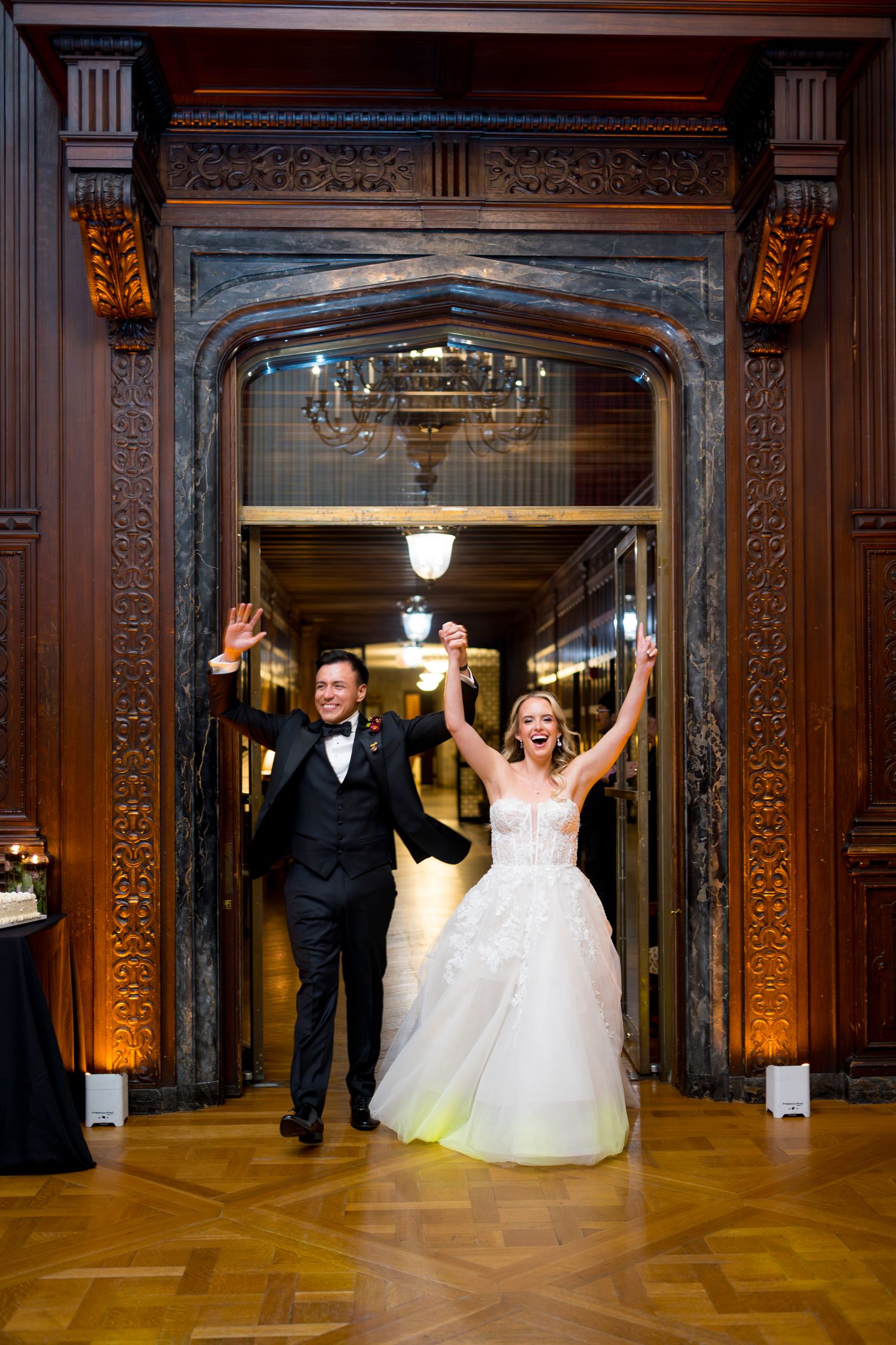 Wedding couple celebrates joyfully in an ornate ballroom with grand wooden decor and parquet flooring.