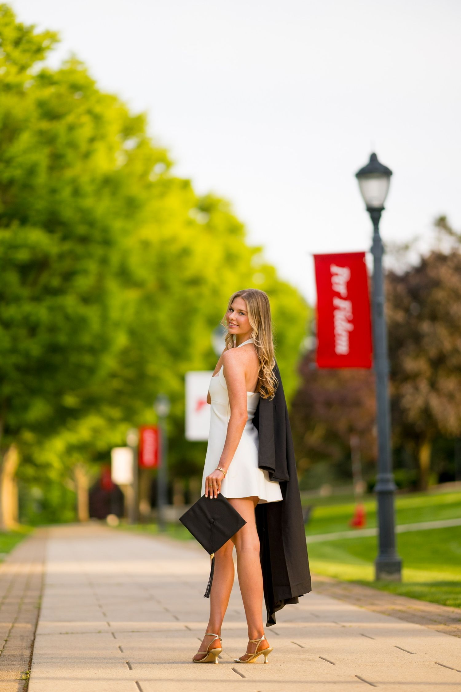 Fairfield University graduate in white dress and black graduation gown poses on campus pathway with red banners.