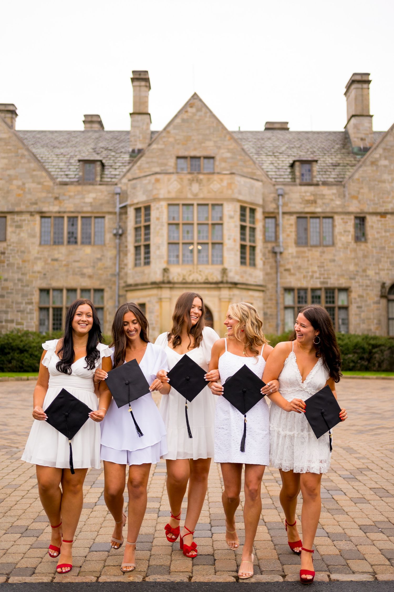 Fairfield University graduates in white dresses hold diplomas while walking together outside stone building.