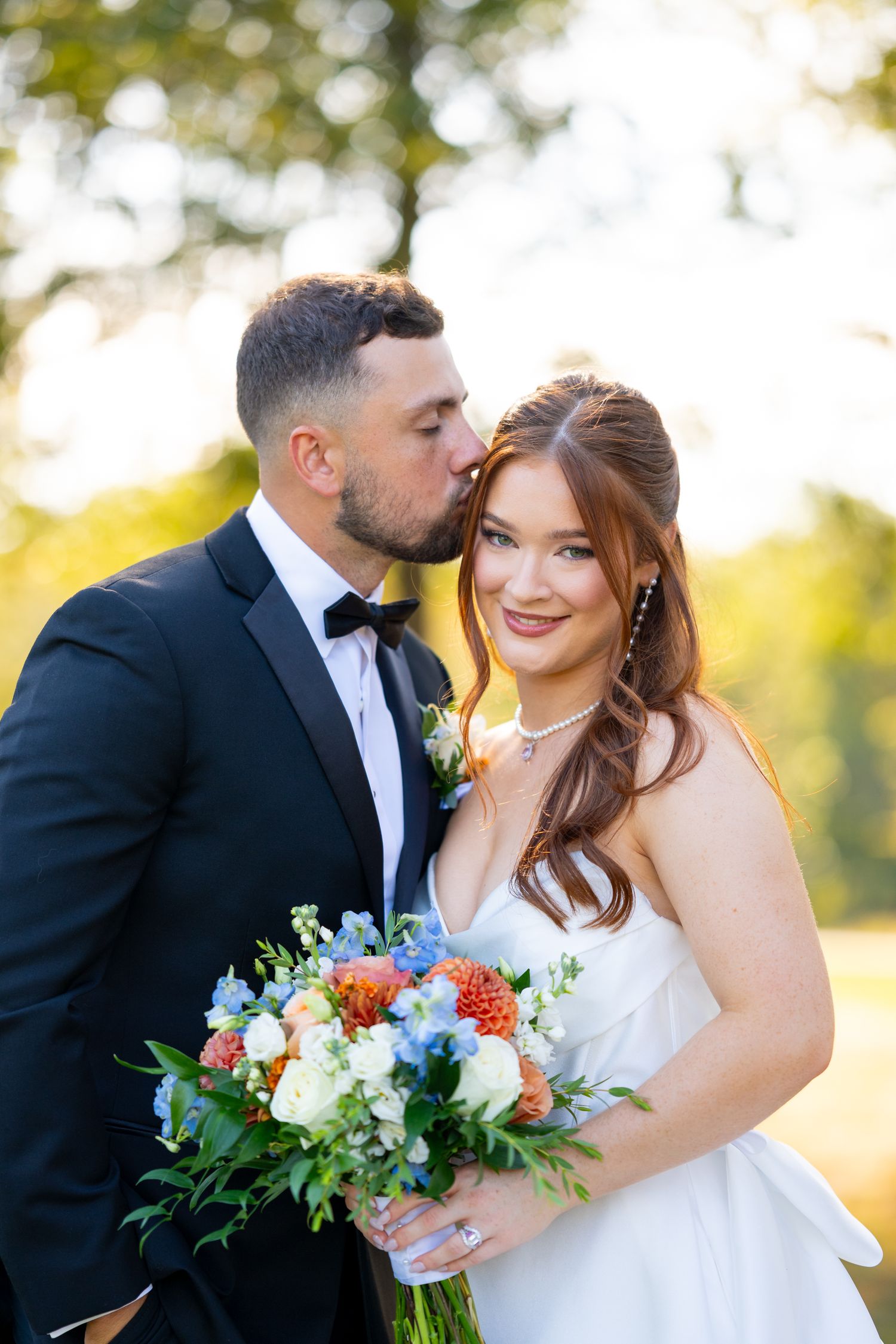 Newlyweds share intimate moments while holding a vibrant blue and orange bouquet at sunset.