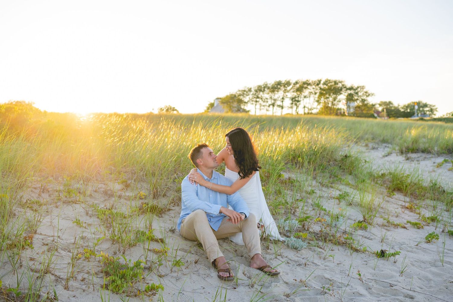 Couple cuddling on sandy Cape Cod beach with ocean waves and sunset in background.