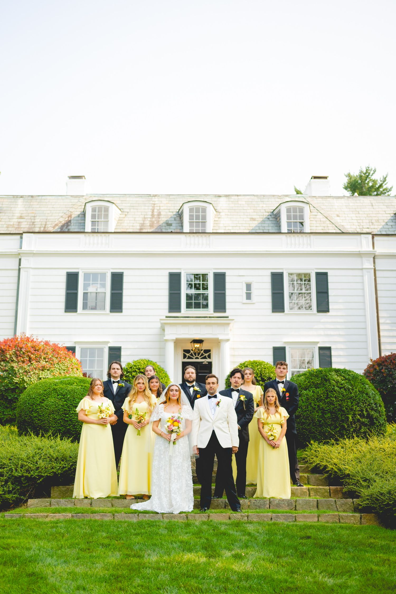 Wedding party in formal attire poses on lawn with white colonial mansion backdrop.