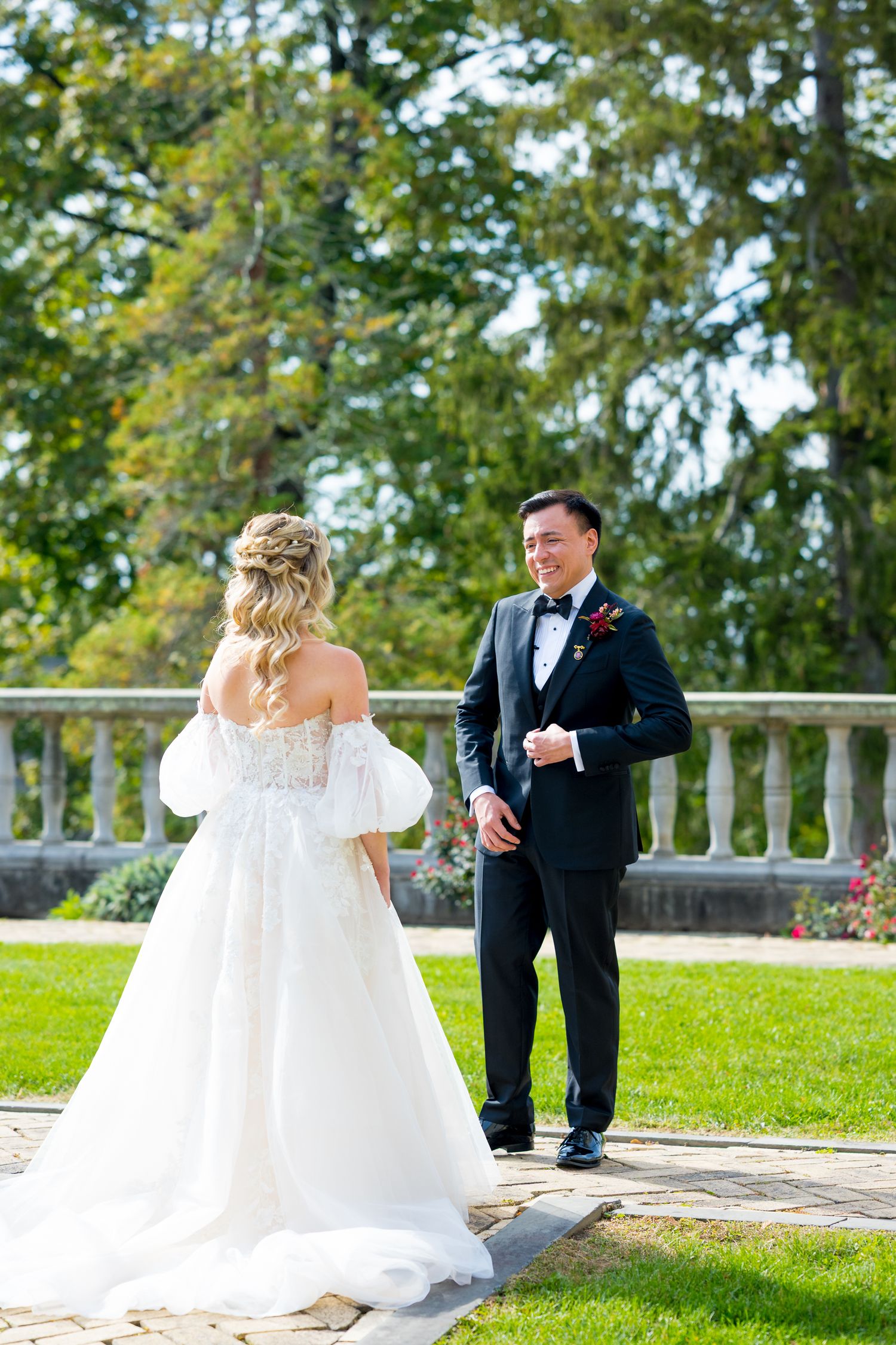 A bride and groom share a joyful first look moment on a sunny terrace surrounded by green trees.