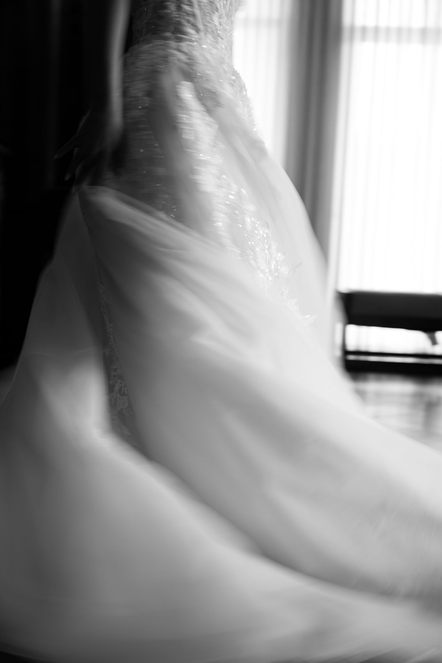 A close-up black and white photograph of an elegant white wedding dress with delicate lace details and flowing fabric.