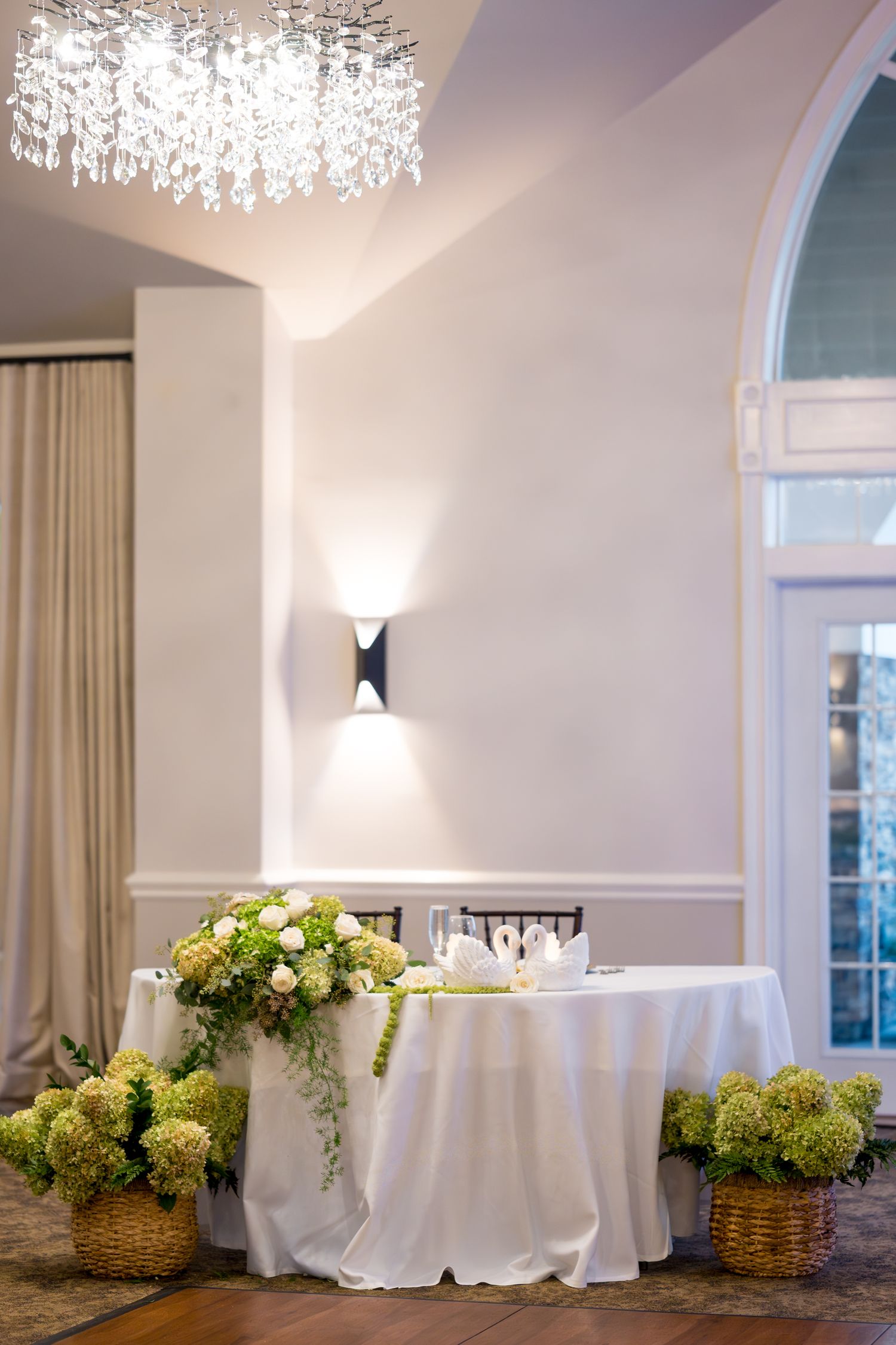 Elegant wedding reception venue with crystal chandeliers, white drapes, and floral arrangements in woven baskets.