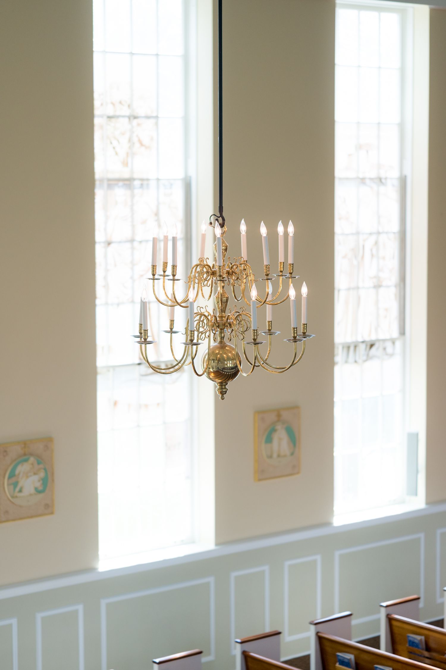 Elegant gold chandeliers hanging in a bright white room with tall windows and wainscoting.
