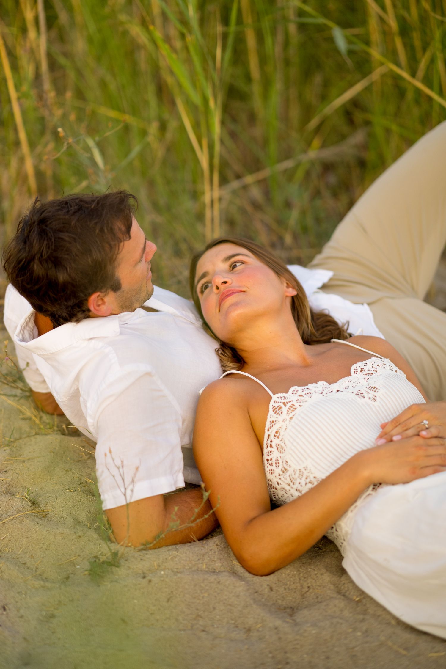 Couple lying together on sandy beach with tall grass in background during sunset.