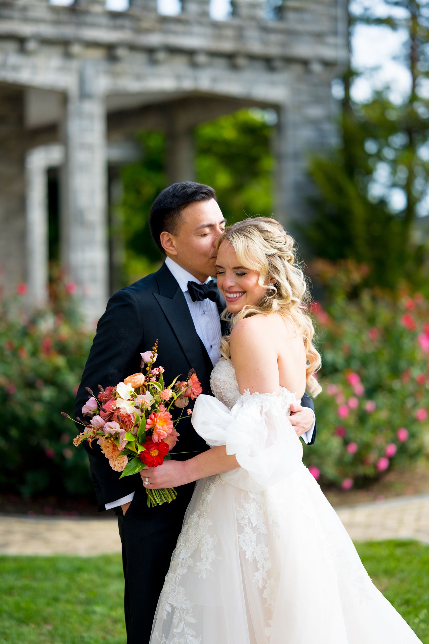 Romantic wedding couple embracing near stone columns and rose garden.