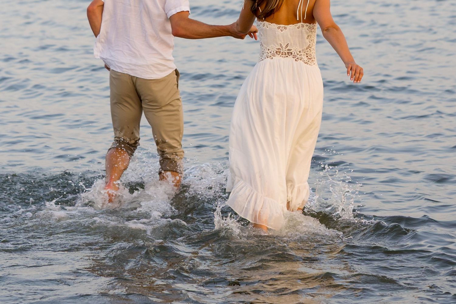 A couple wades through shallow ocean water, with the woman wearing a flowing white dress and lace details.