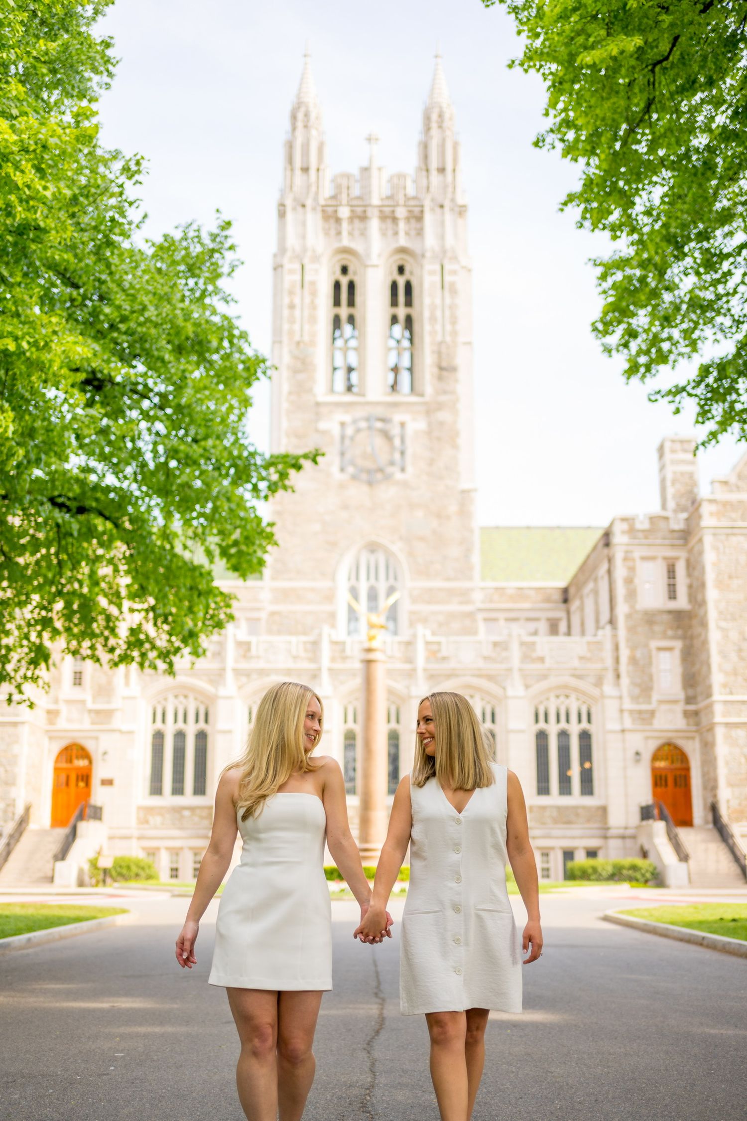 Two Boston College graduates in white dresses walking in front of university building.