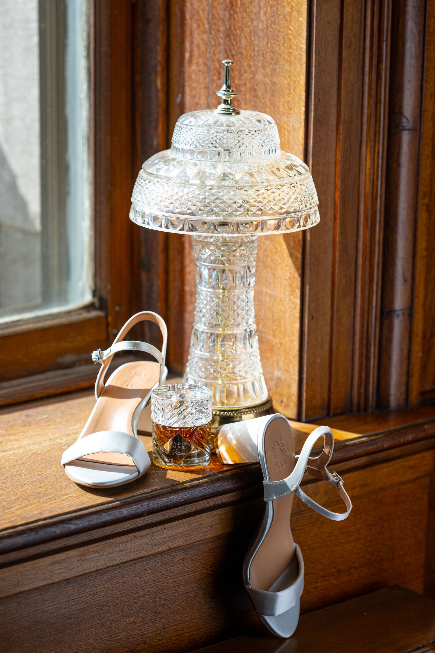 White bridal shoes and perfume displayed next to crystal decorative lamp on a wooden windowsill.