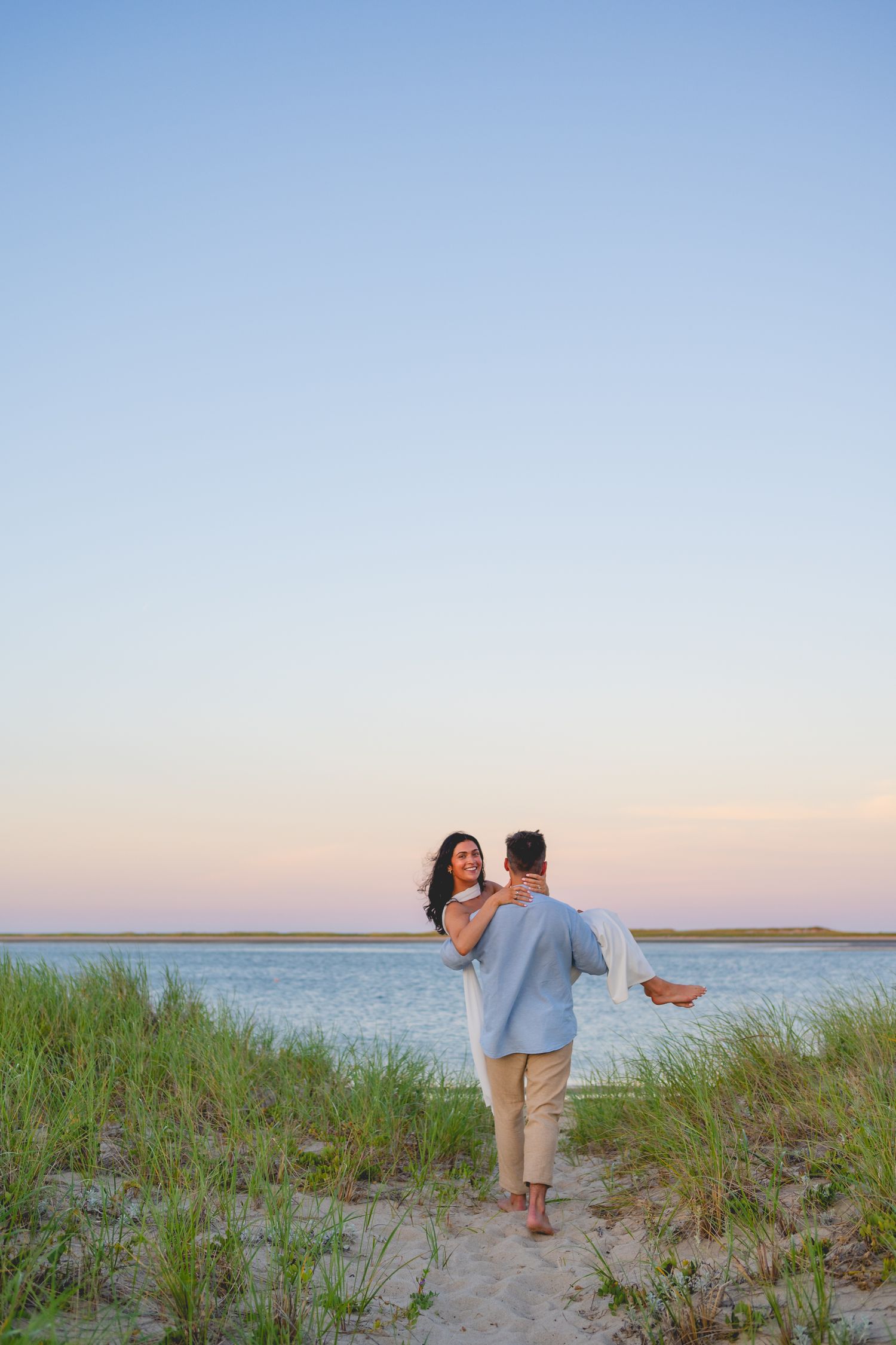 Couple walking through beach grass and dunes with Cape Cod ocean view at sunset.