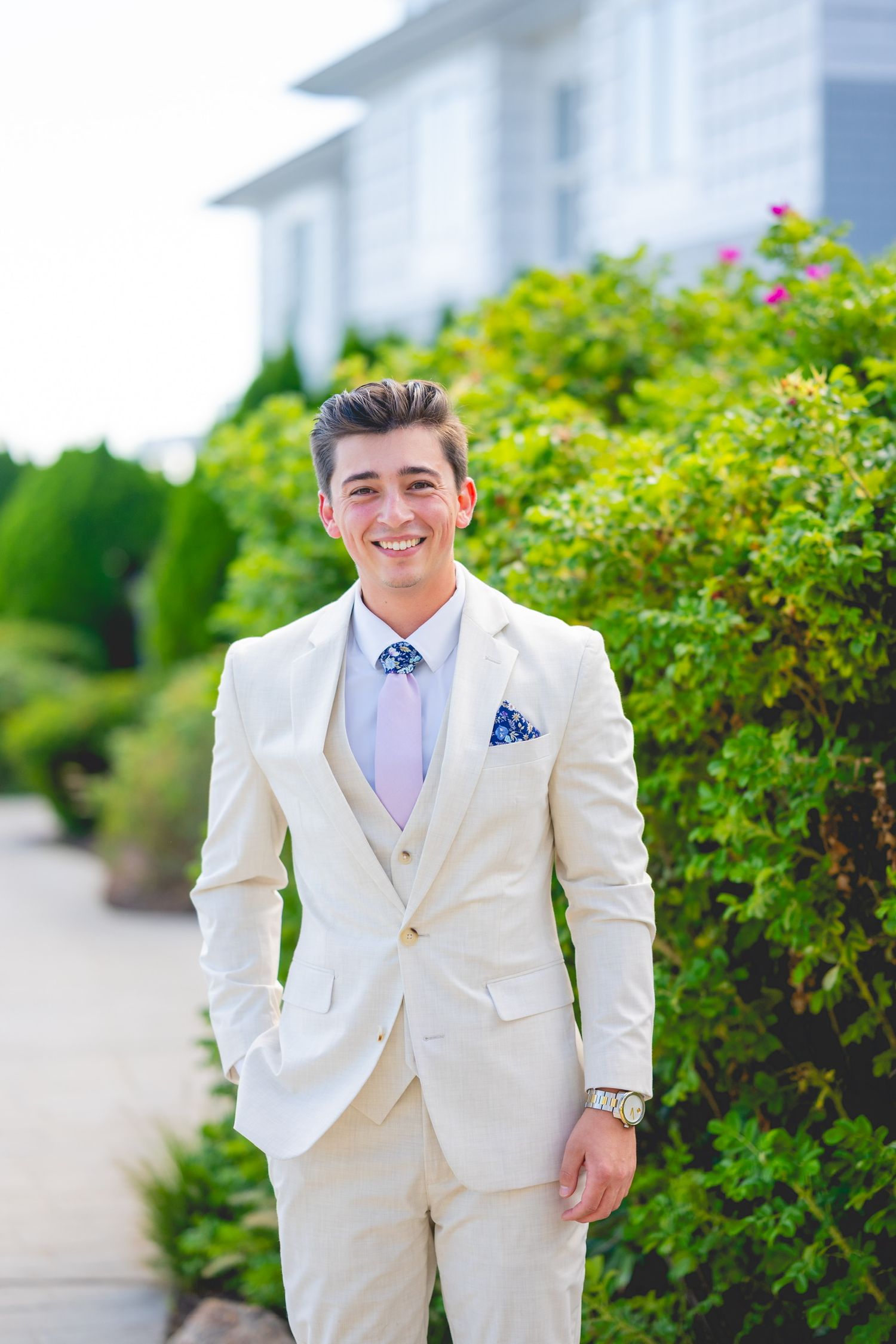 Groom in crisp white tuxedo smiles while standing by garden greenery at Water's Edge Resort & Spa.