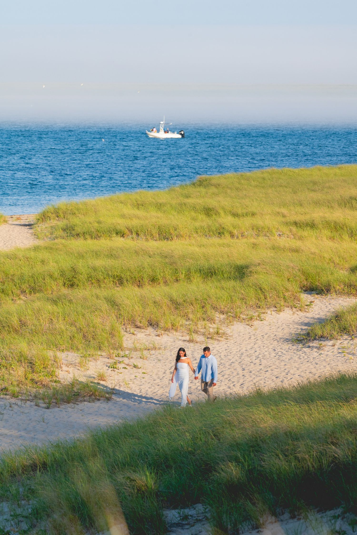 Coastal Cape Cod wedding photos with couple walking along beach path with ocean and grassy dunes.
