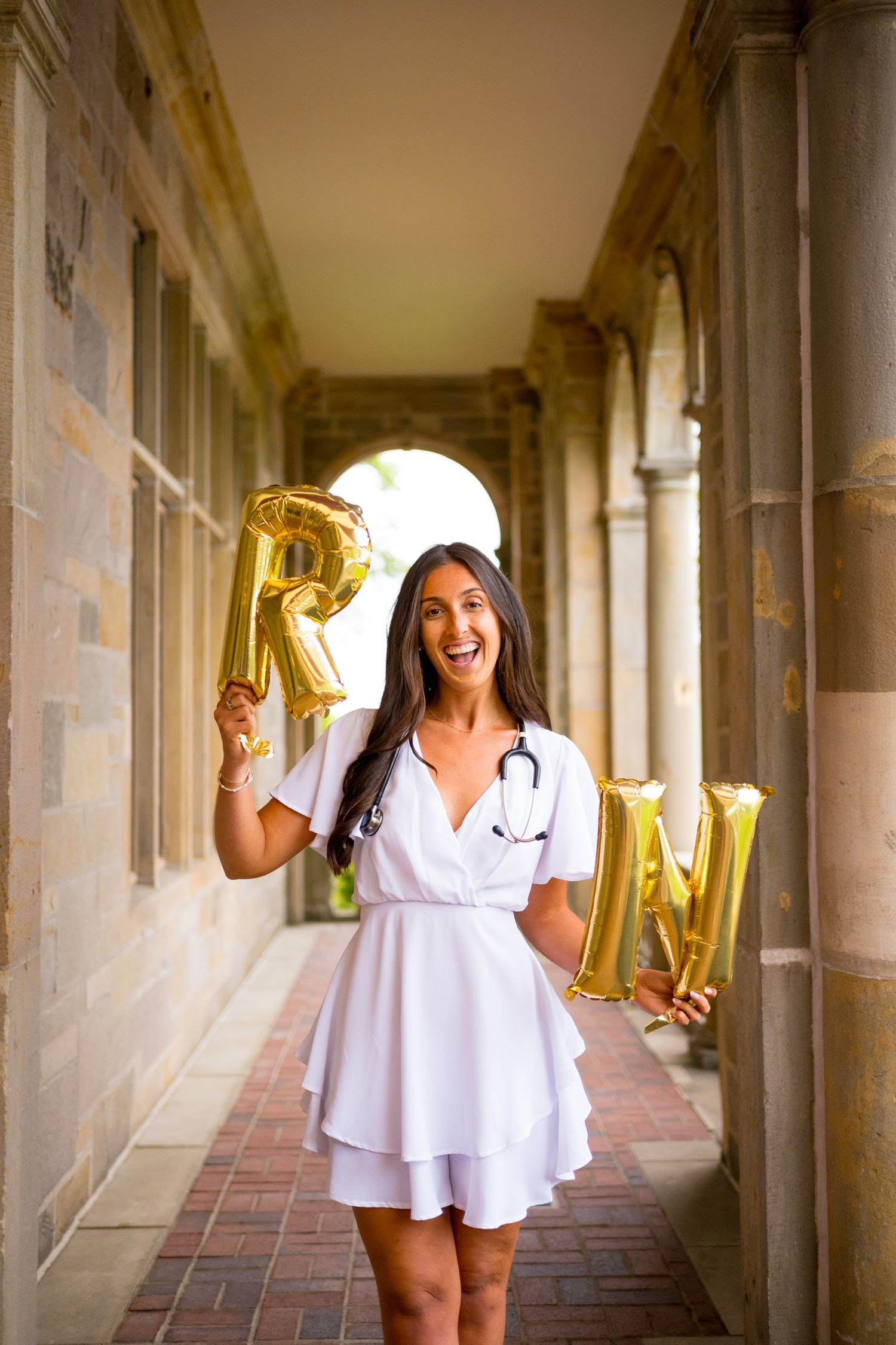 Fairfield University graduate in white dress holds gold letter balloons in stone hallway.