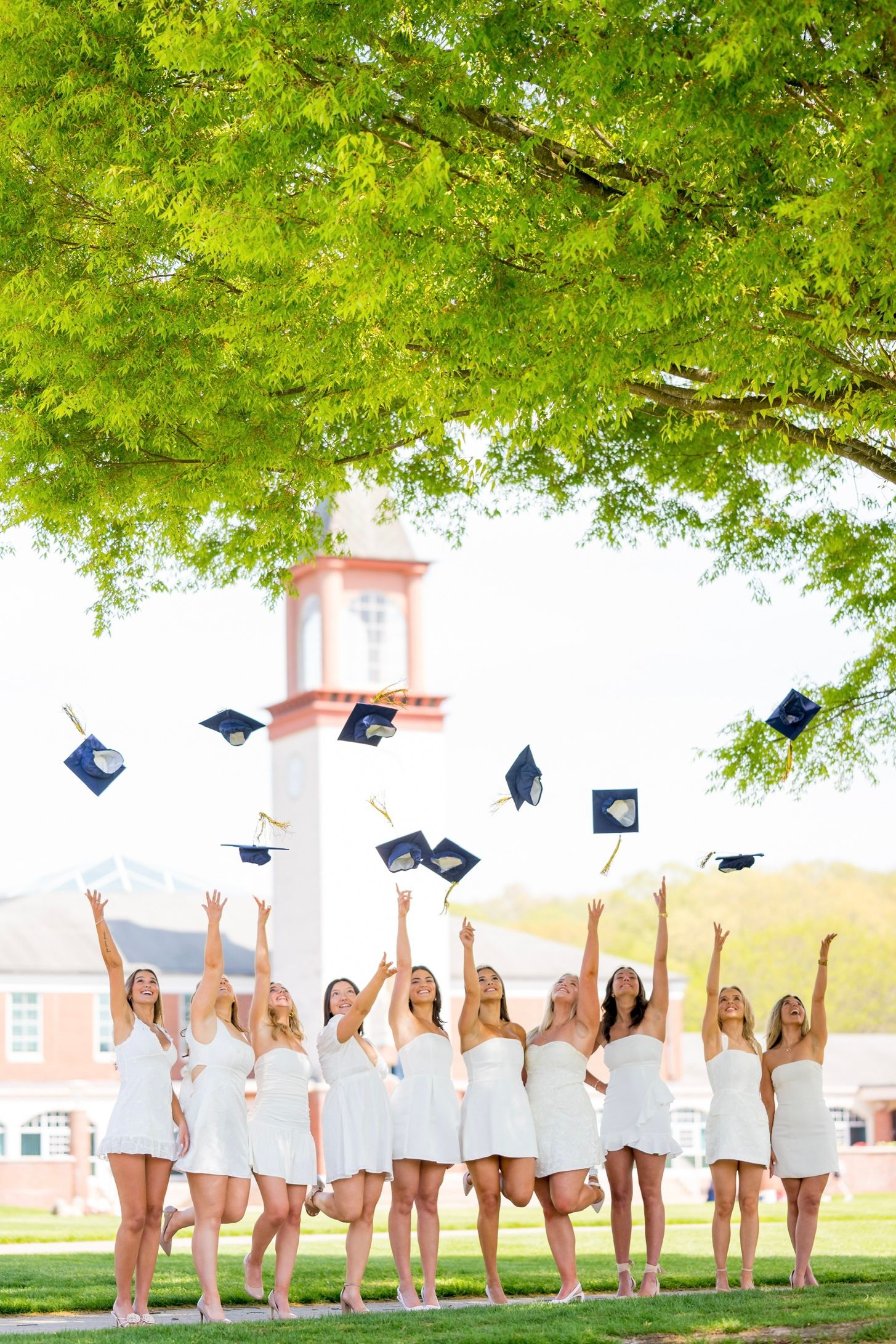 Quinnipiac University graduates in white dresses toss their graduation caps into the air under green trees.