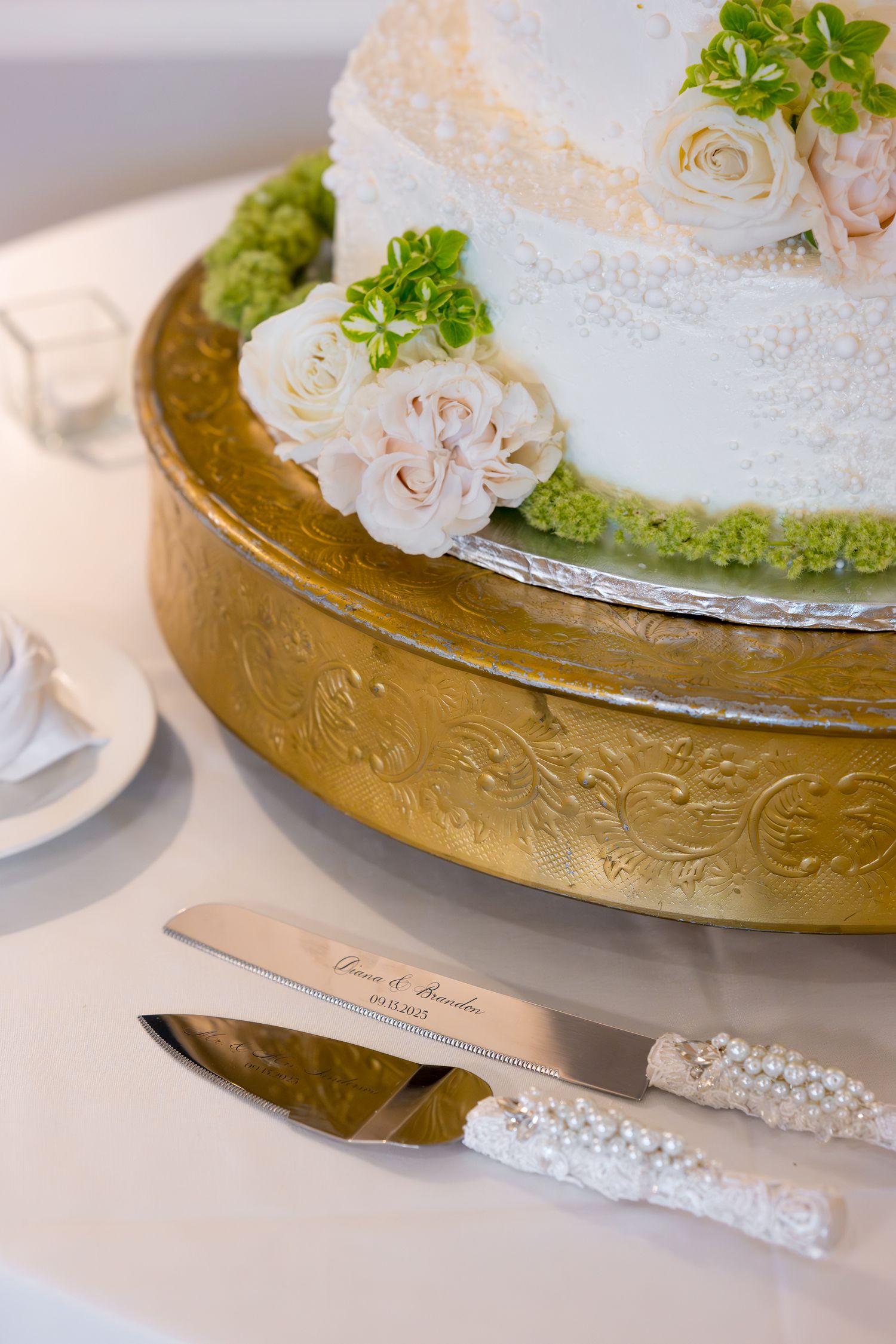 White wedding cake with blush roses and greenery sits on ornate gold cake stand with pearl-handled serving set.