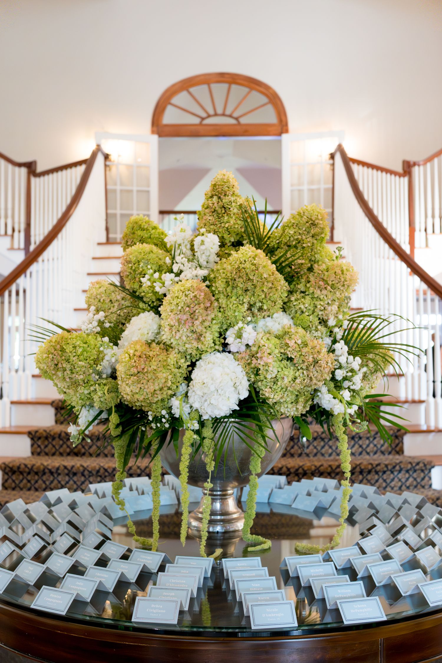 Elegant floral arrangement of green hydrangeas and white flowers sits atop place card display at wedding venue entrance.