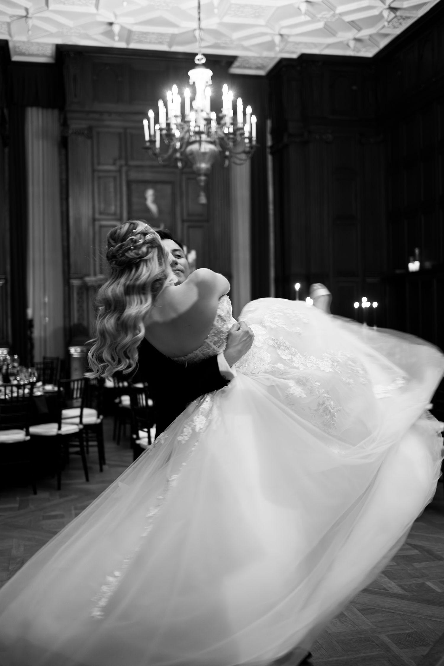 A bride in a flowing white ballgown spins gracefully on a grand ballroom floor under chandeliers.