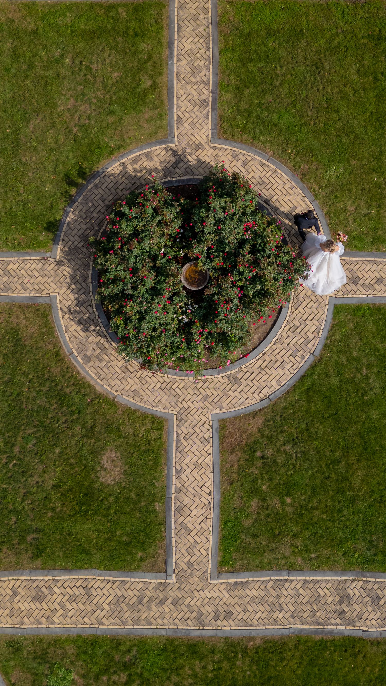 Aerial view of a circular garden with paved pathways and a couple in formal attire standing at the edge.