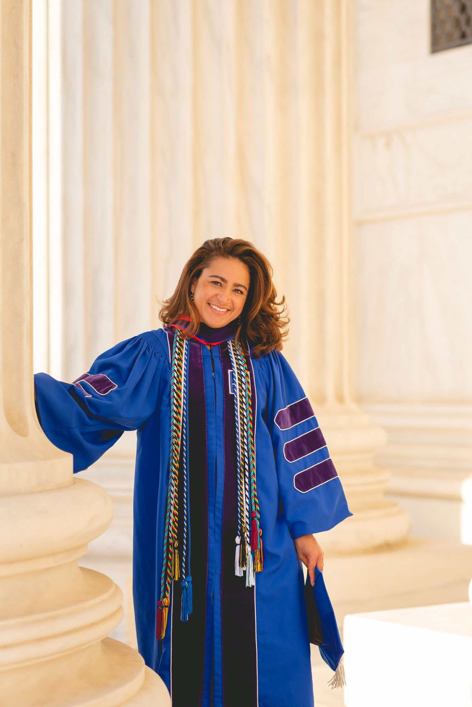 Graduate in blue academic regalia poses playfully against white columned background in Washington D.C. for graduation photos.