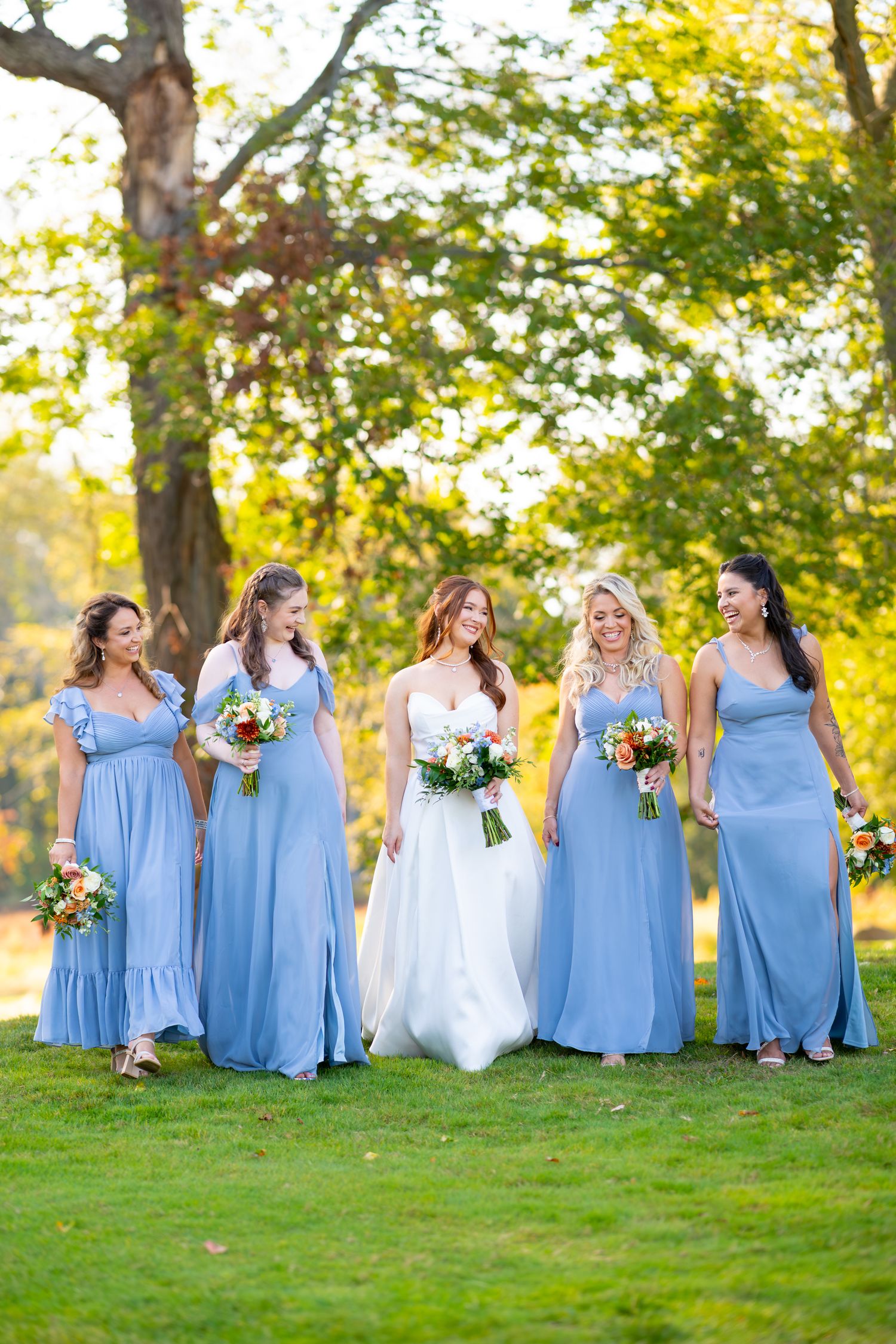 Wedding party in light blue bridesmaid dresses poses on sunny lawn with bouquets.