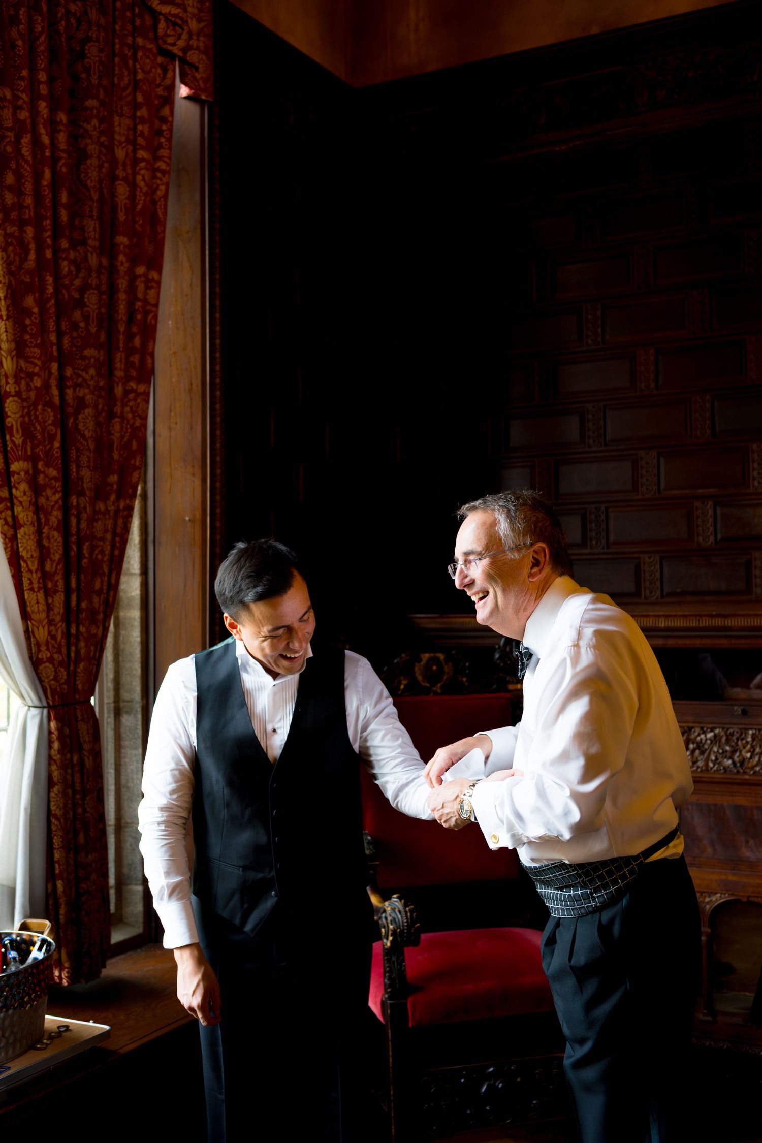 Two people in formal attire share a candid moment in a room with red curtains and dramatic lighting.