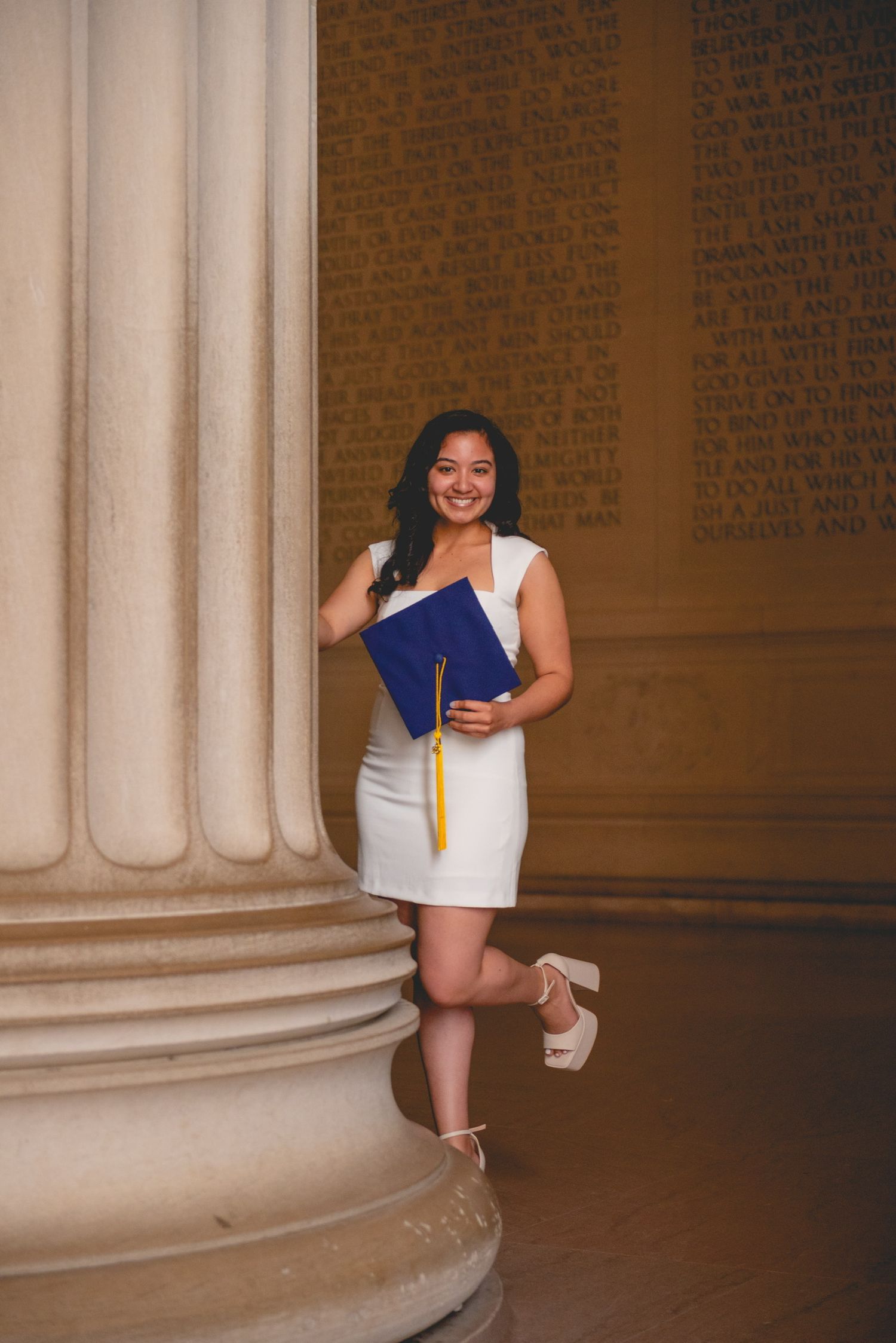 Graduate in white dress poses playfully around classical stone columns during photo shoot.