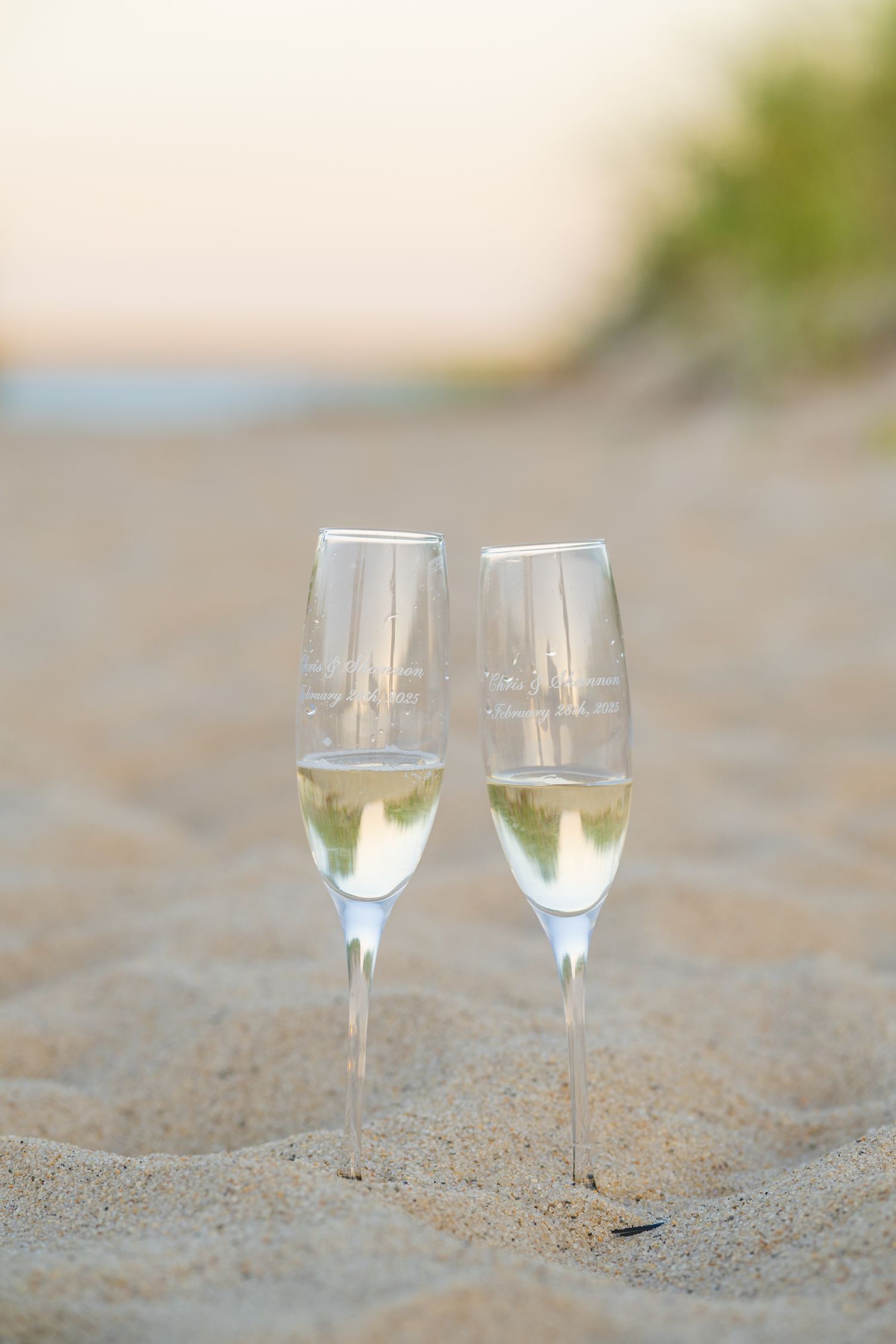 Two champagne flutes with sparkling wine sit nestled in sandy beach at sunset.