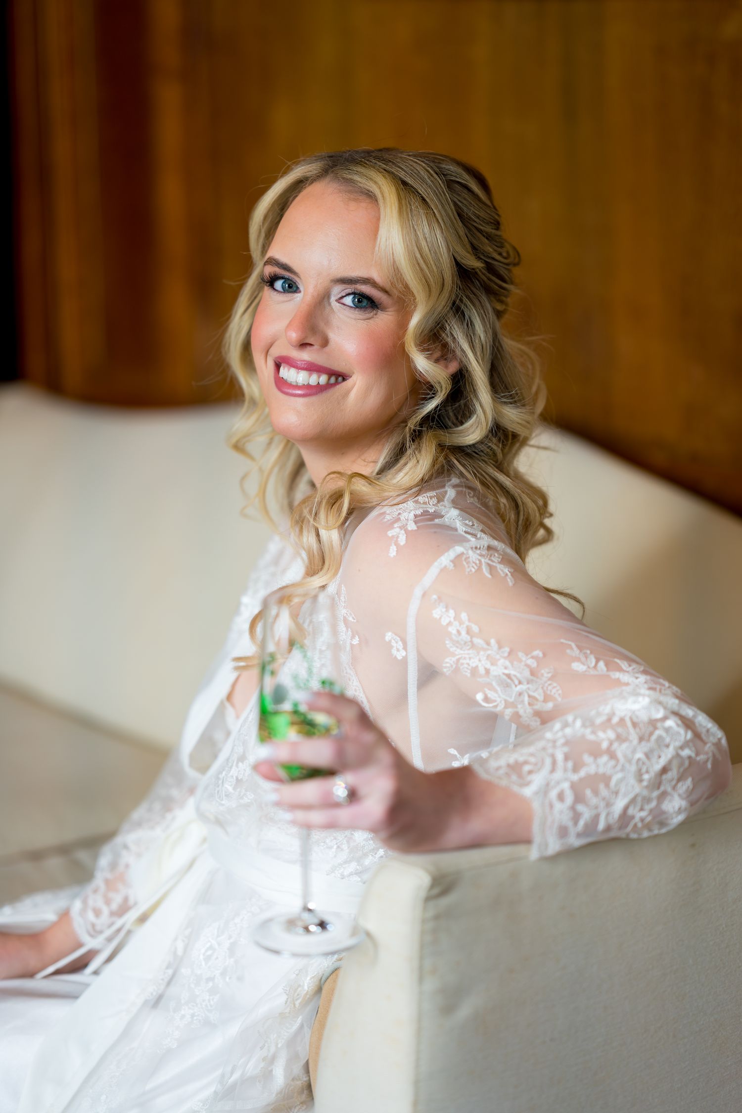 A bride in a lace wedding dress holds a champagne glass while getting ready in a warm-lit room.