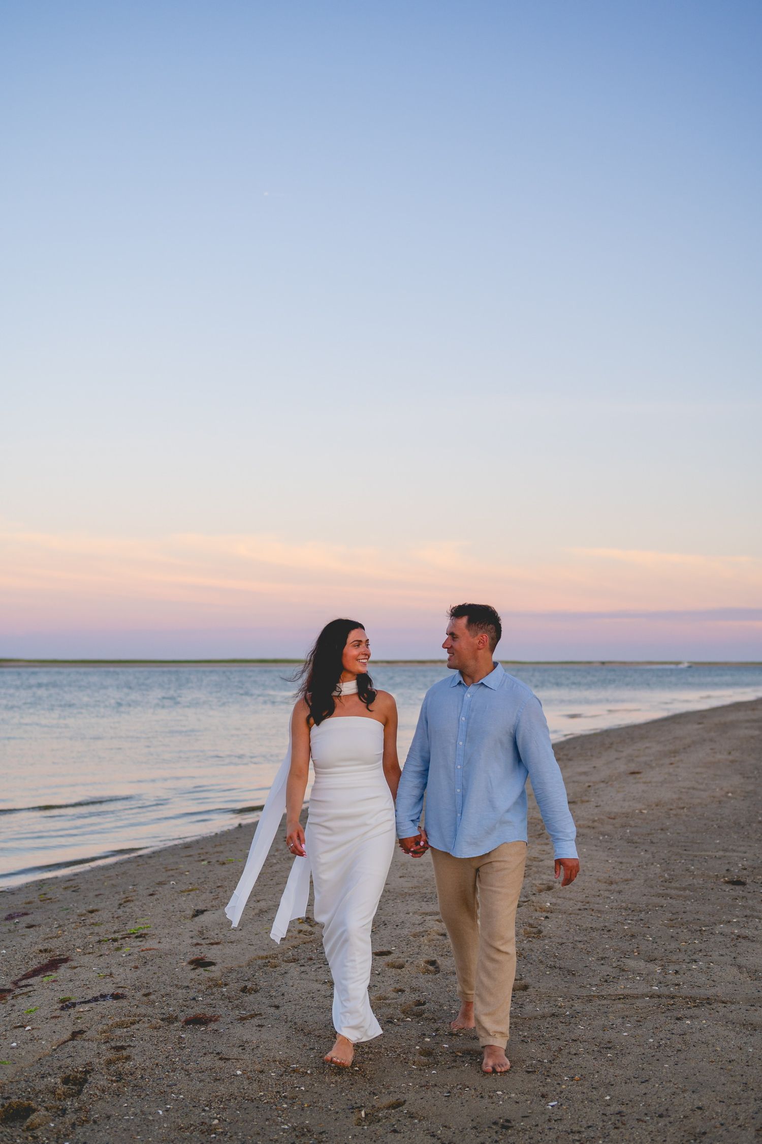 Couple walking together on a peaceful Cape Cod beach at sunset wearing white and khaki casual attire.
