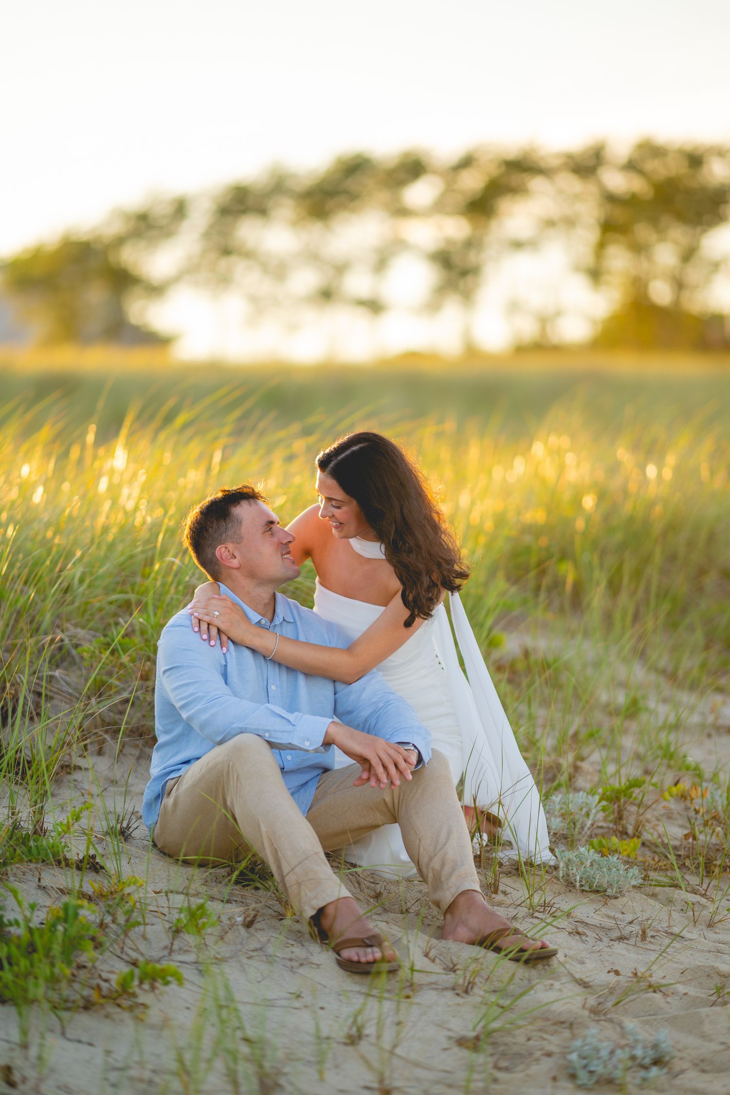 A couple sits together on sandy dunes during golden hour at Chatham Lighthouse Beach in Chatham, Massachusetts.