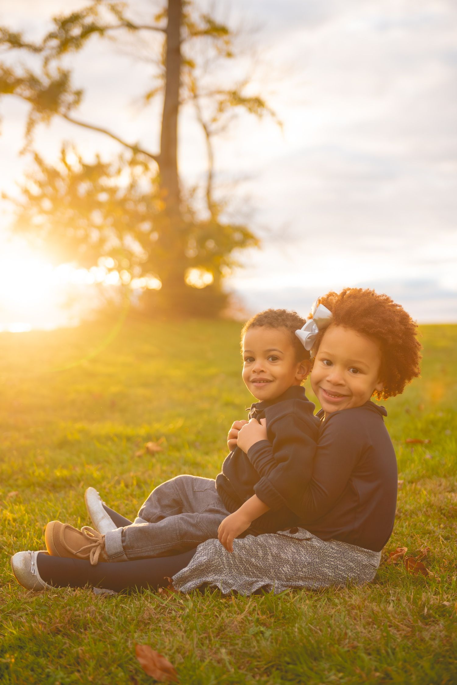 Two young children sit together on grass during golden hour sunset.