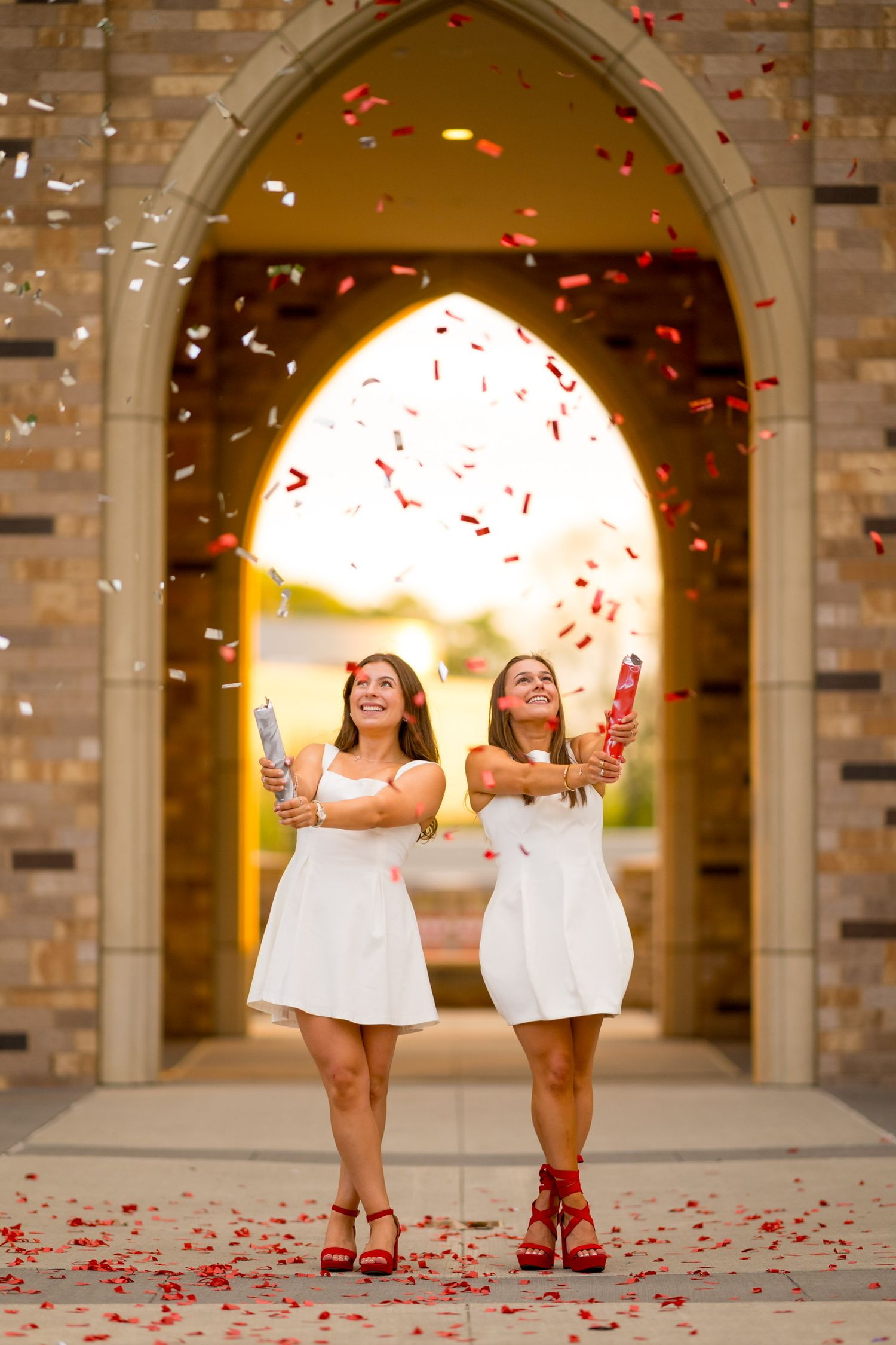 Two Sacred Heart University graduates in white dresses throw rose petals under illuminated stone archway at sunset.