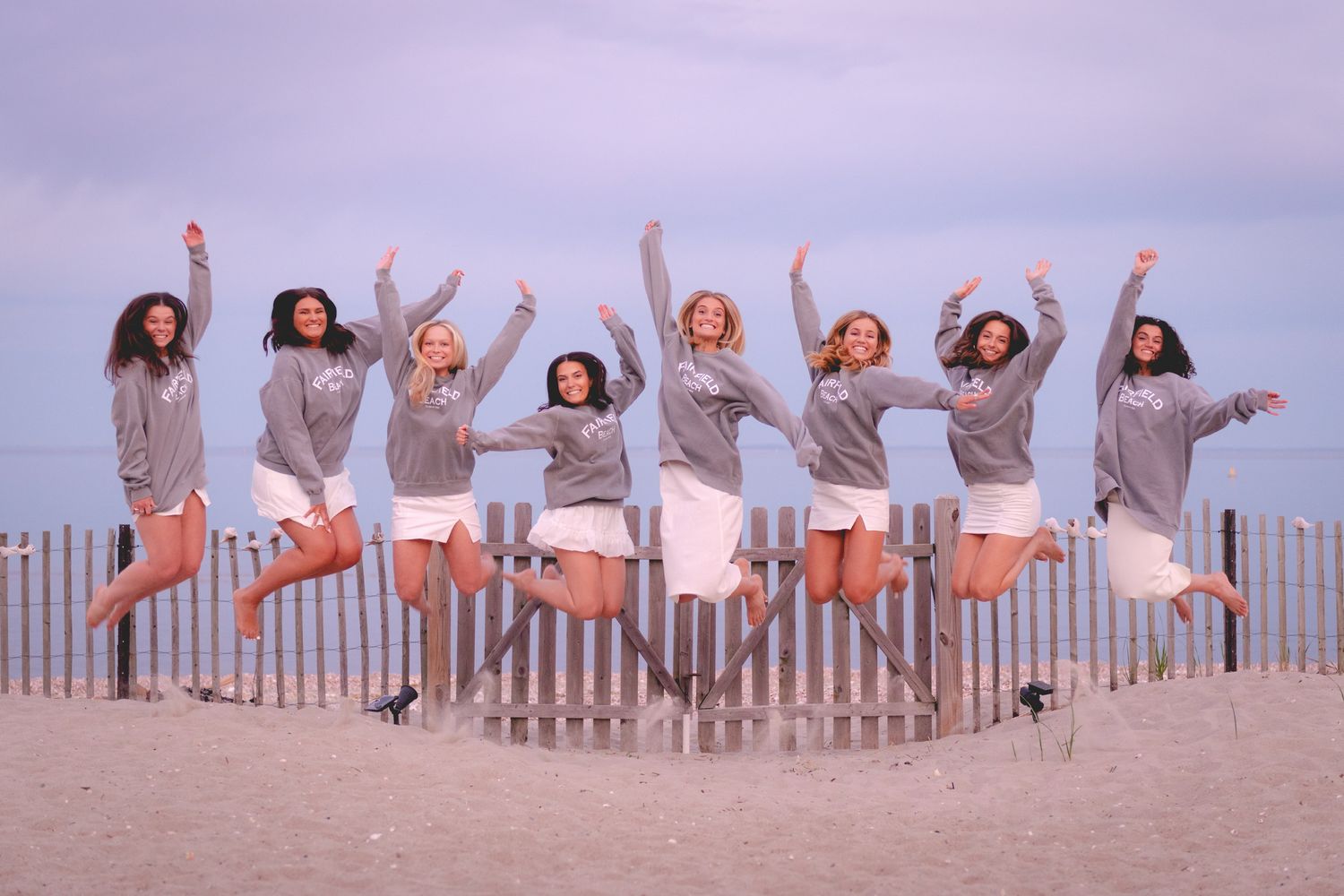 Group of friends in matching outfits jumping joyfully on beach during sunset celebration.