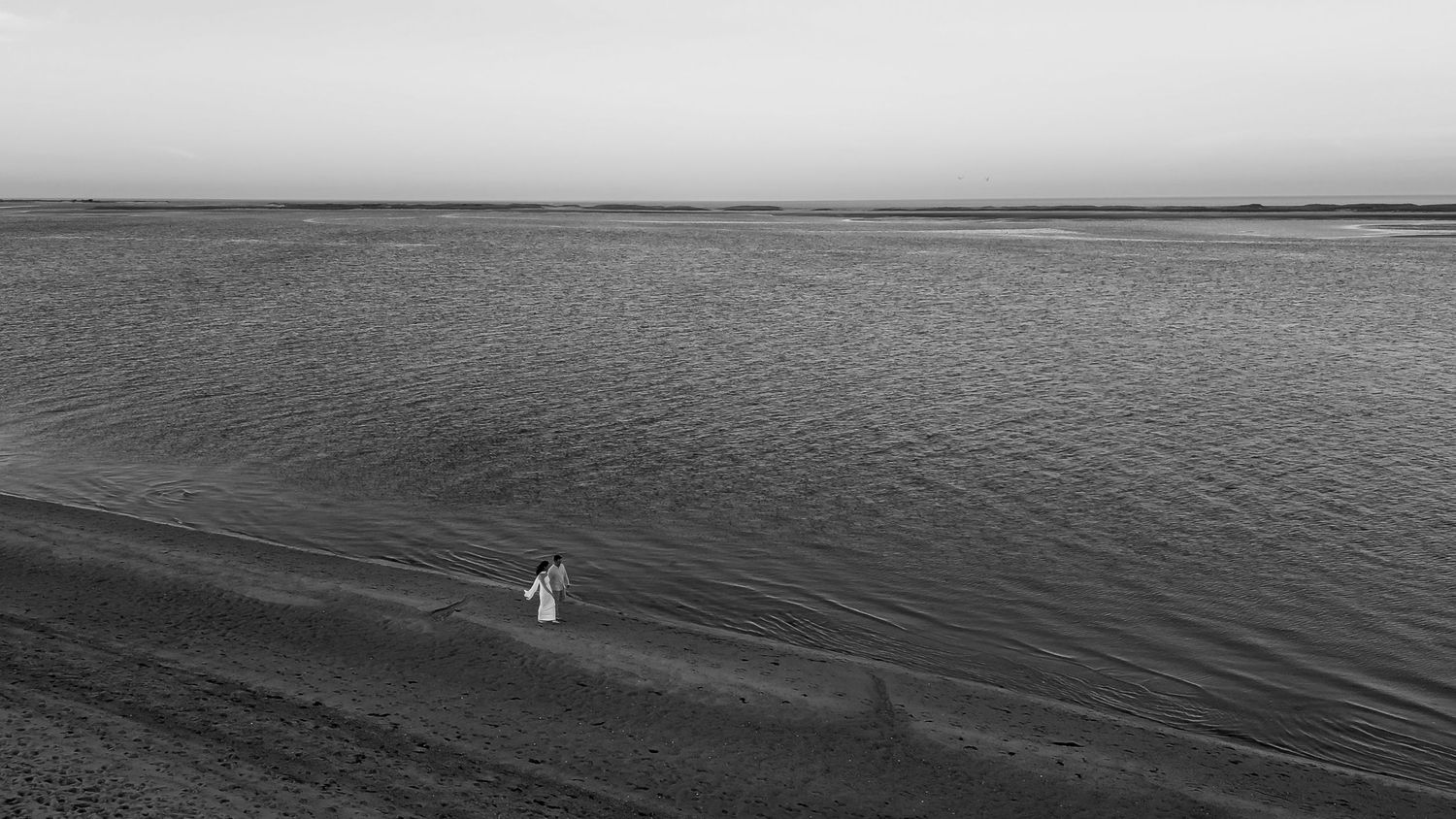 A solitary figure in white stands on a dark shoreline against vast waters in a minimalist black and white landscape.