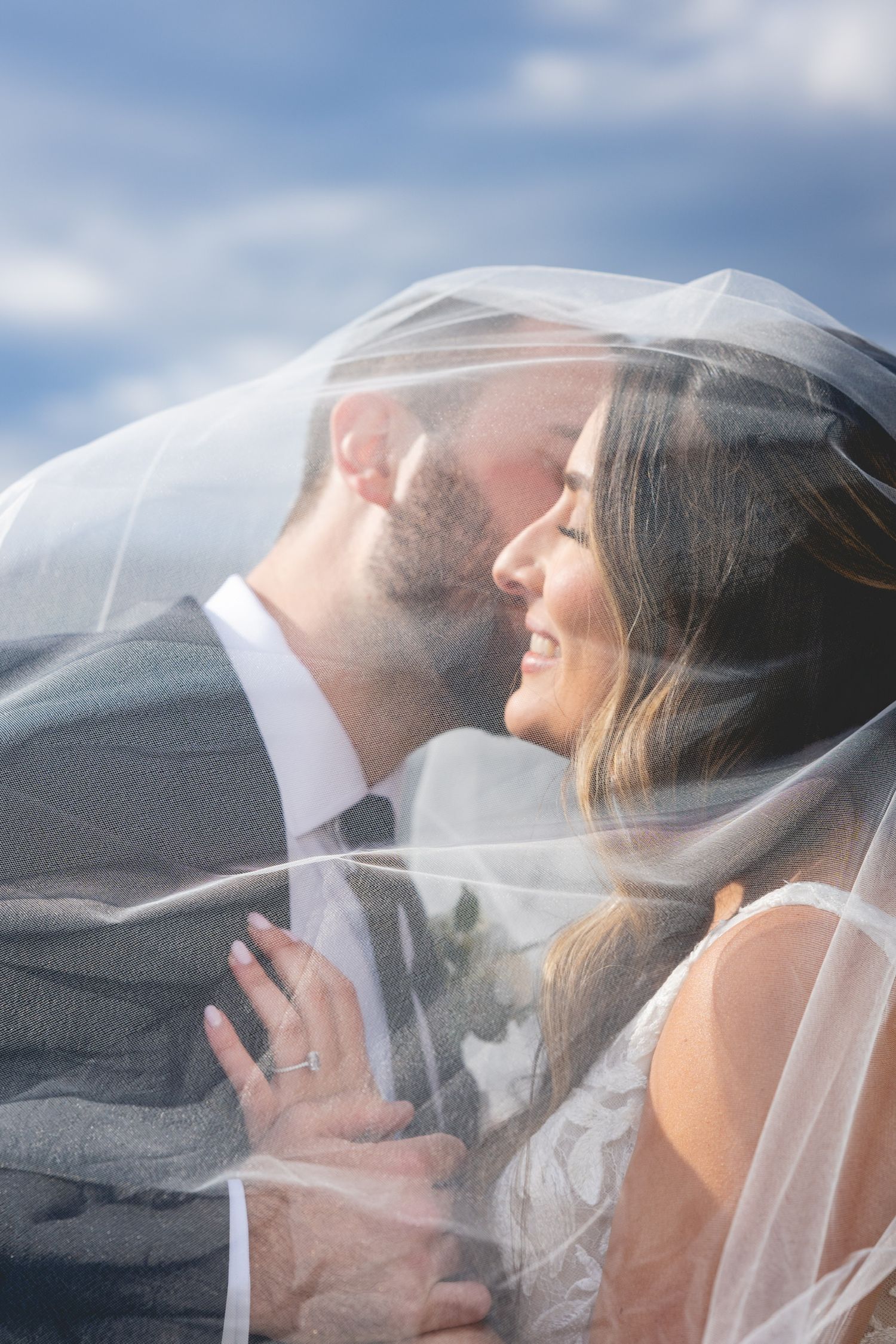 Romantic wedding couple sharing an intimate moment under a flowing veil against a blue sky background.