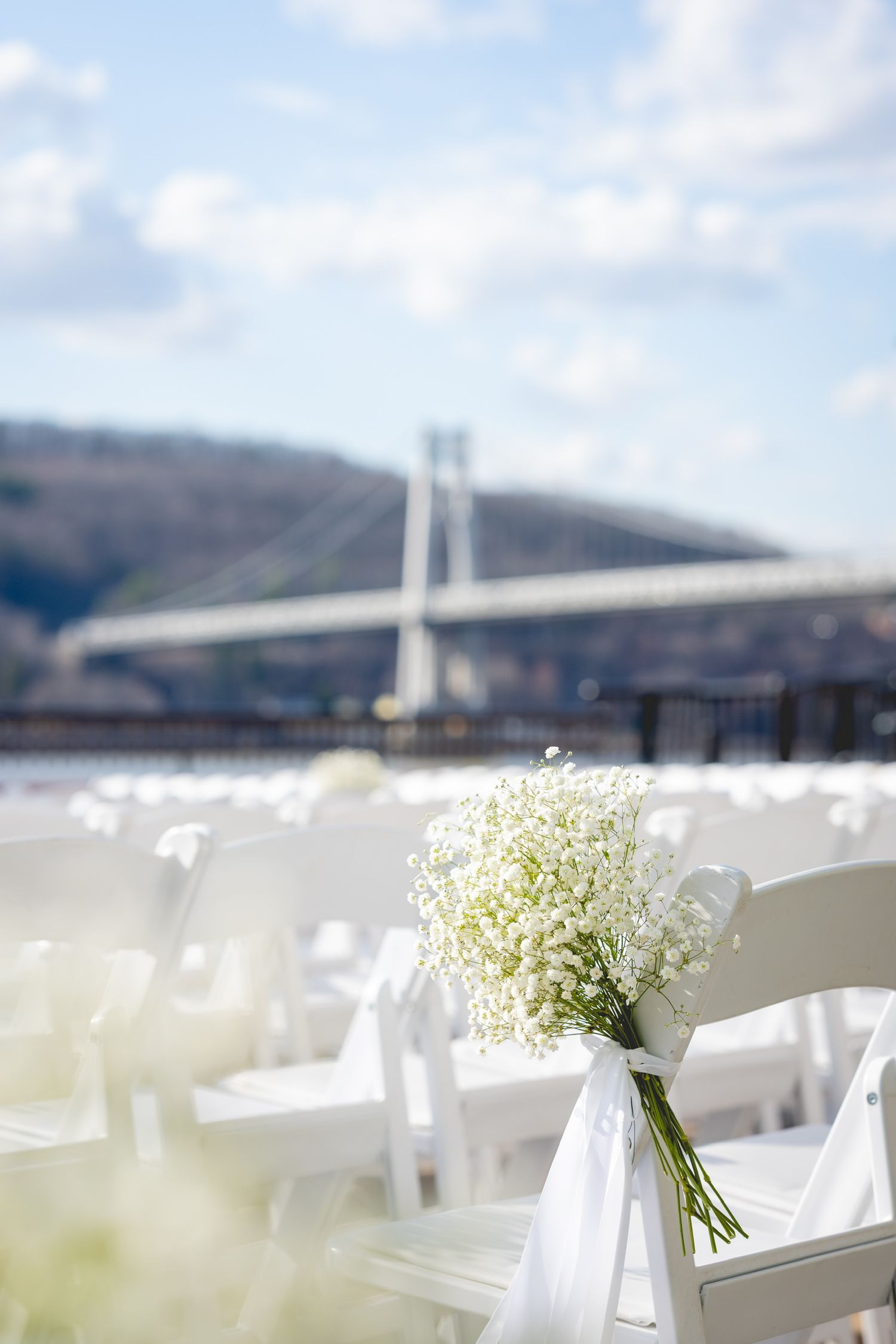 White folding chairs with baby's breath bouquets line up with a bridge view in the background.