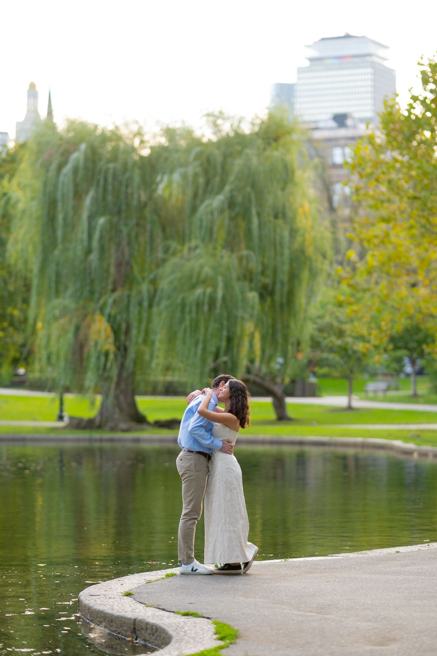 A couple embraces by a pond with weeping willows in a scenic Boston Public Gardens setting.