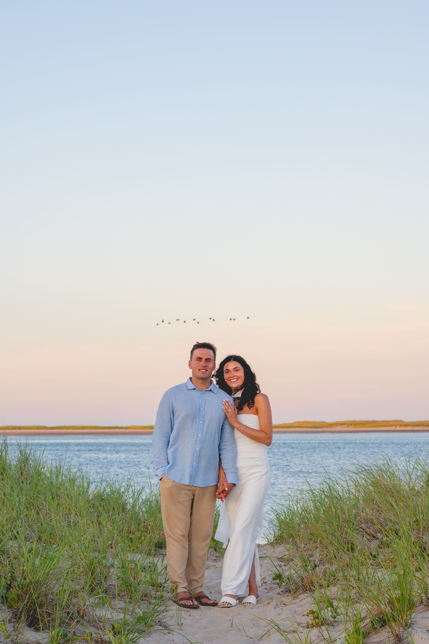 A couple walks along a scenic beach in Cape Cod at sunset with tall grass and ocean views.